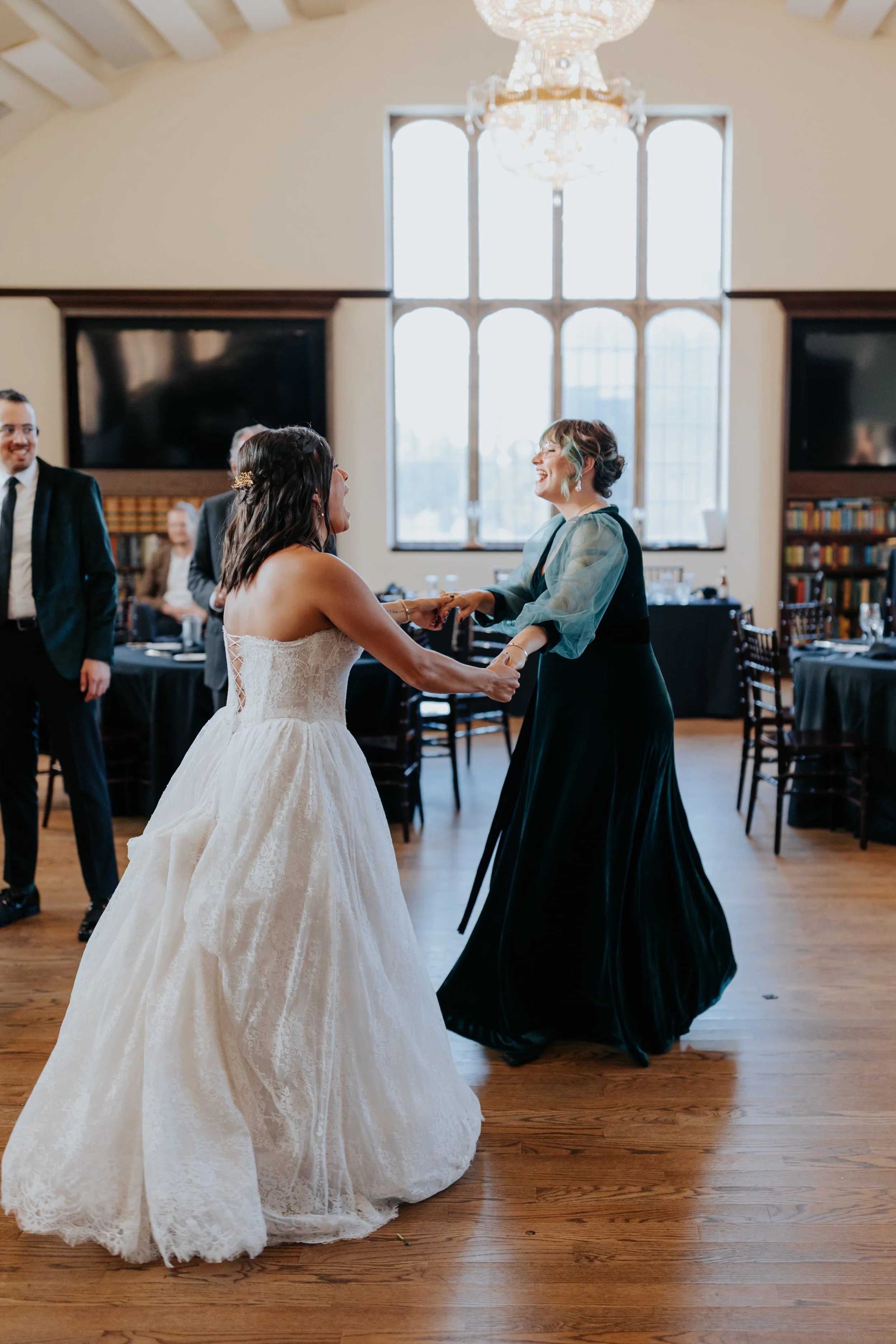 Two women dancing, one in a white wedding dress and the other in a dark dress, at a wedding reception in a banquet hall with large windows and a chandelier.