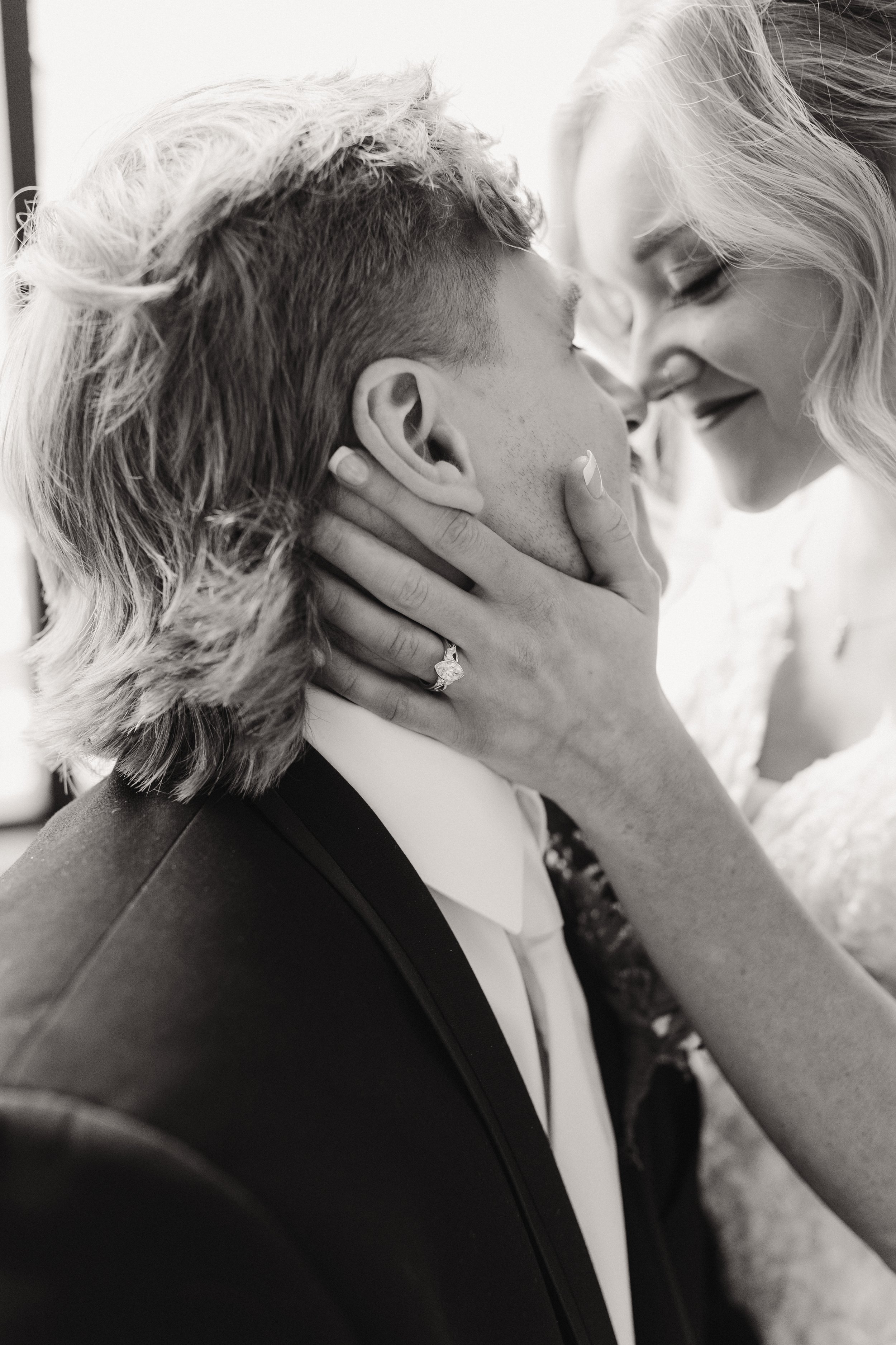 A couple shares a kiss, with the woman holding the man's face, showcasing her engagement ring, in a close-up black-and-white photo.