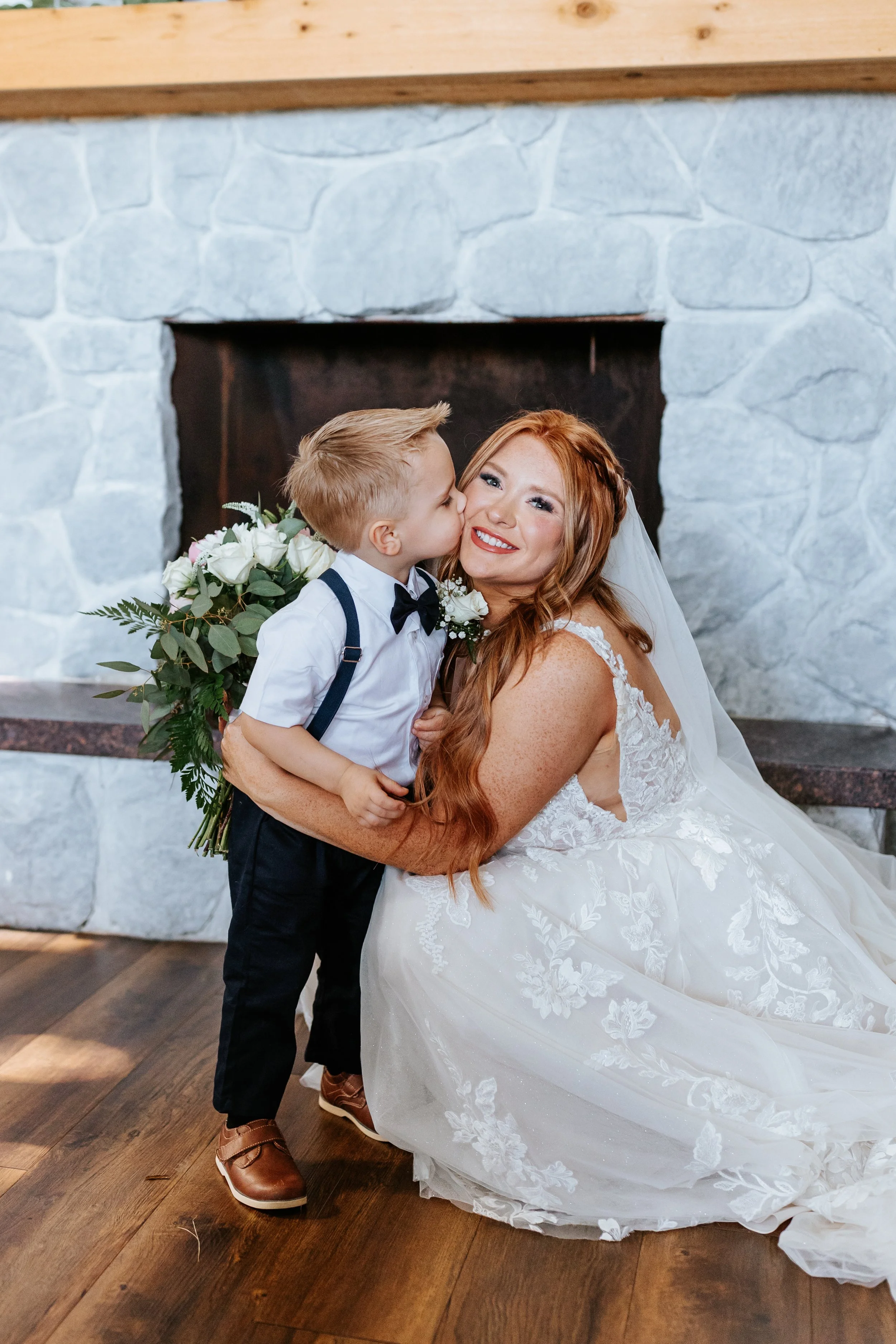 A bride in a white wedding dress, sitting on the floor, is smiling as a young boy kisses her cheek. The boy is dressed in a white shirt, black pants, suspenders, and a bow tie, and holds a bouquet of white flowers with green foliage.