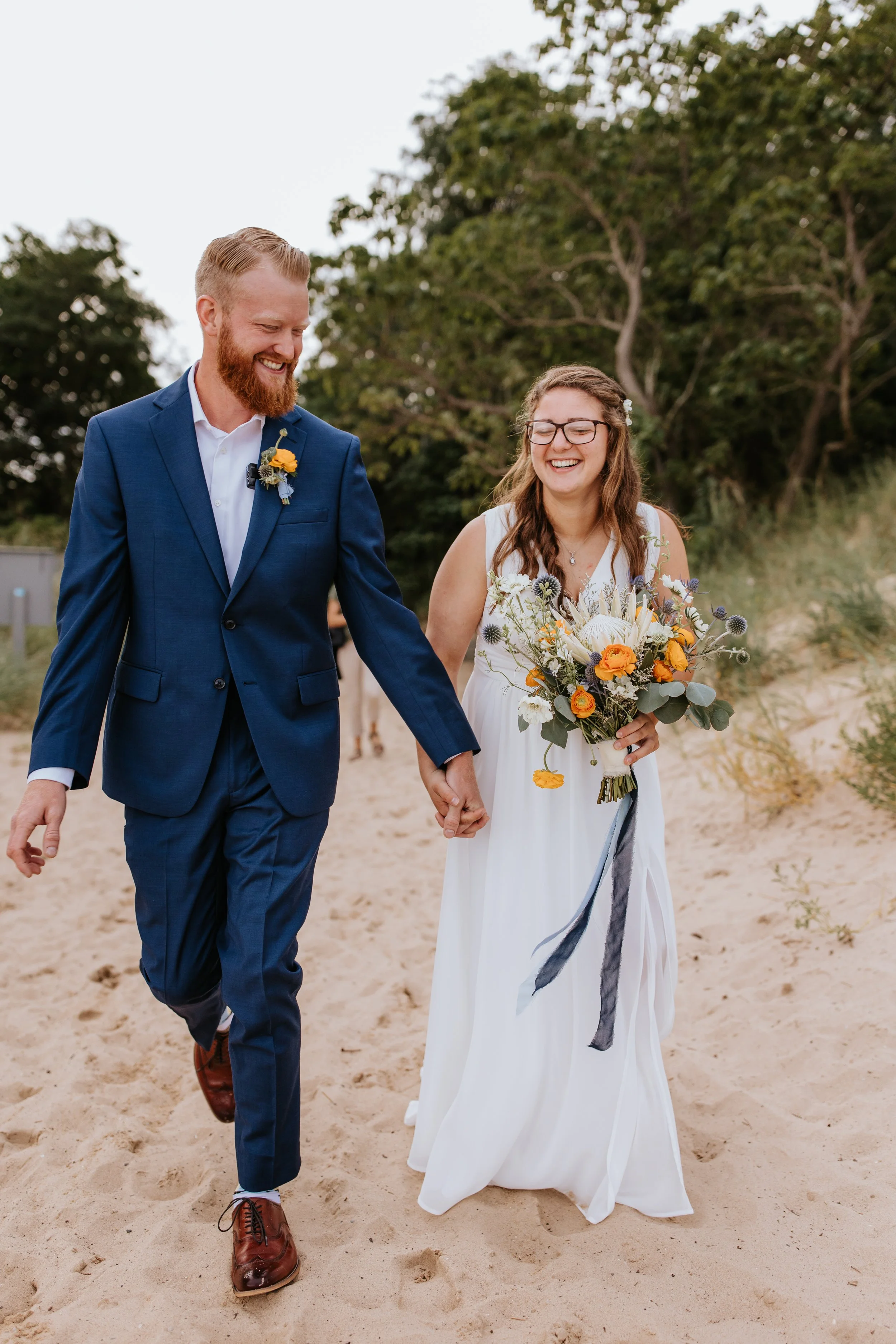 A happy couple walking on the beach, holding hands, with the woman holding a bouquet of flowers, both smiling and dressed in wedding attire.