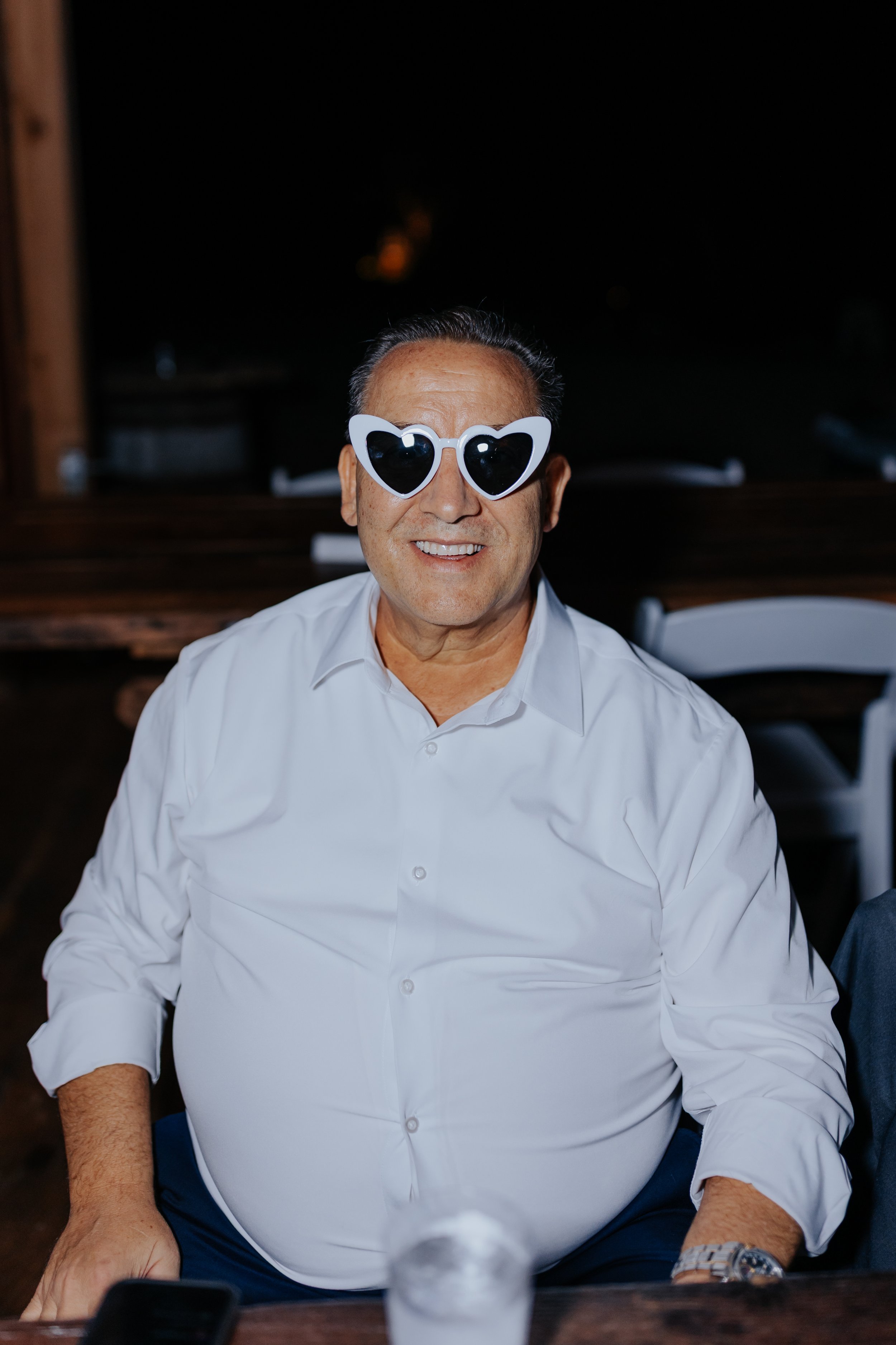 A smiling man with dark hair, wearing a white dress shirt and heart-shaped sunglasses, sitting at a table during an evening event.