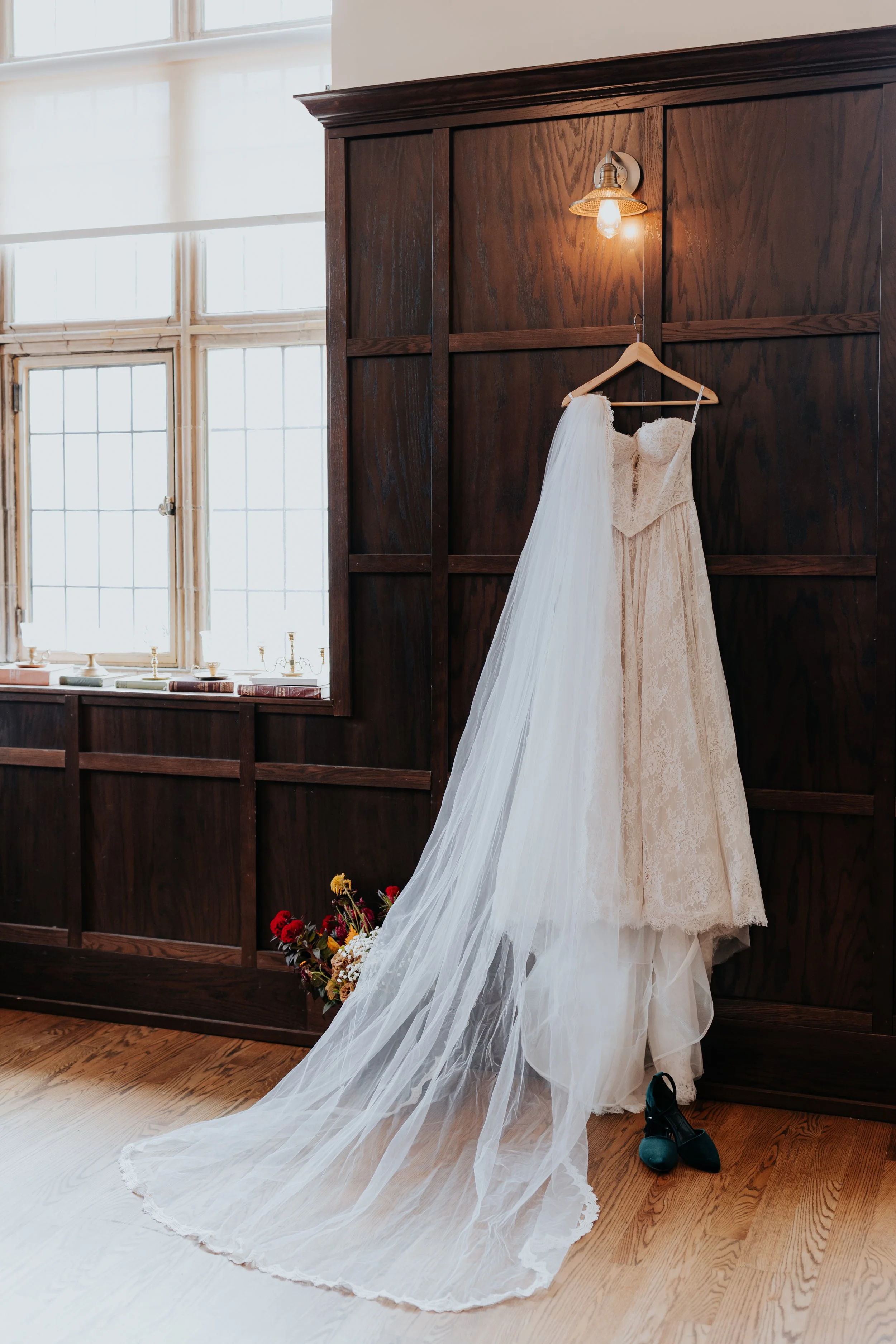 A wedding dress with a long veil hanging on a wooden hanger on a dark wood-paneled wall. A small light fixture is above the dress. Below the dress, there are black shoes and a bouquet of flowers on a wooden floor near a large window with white frames