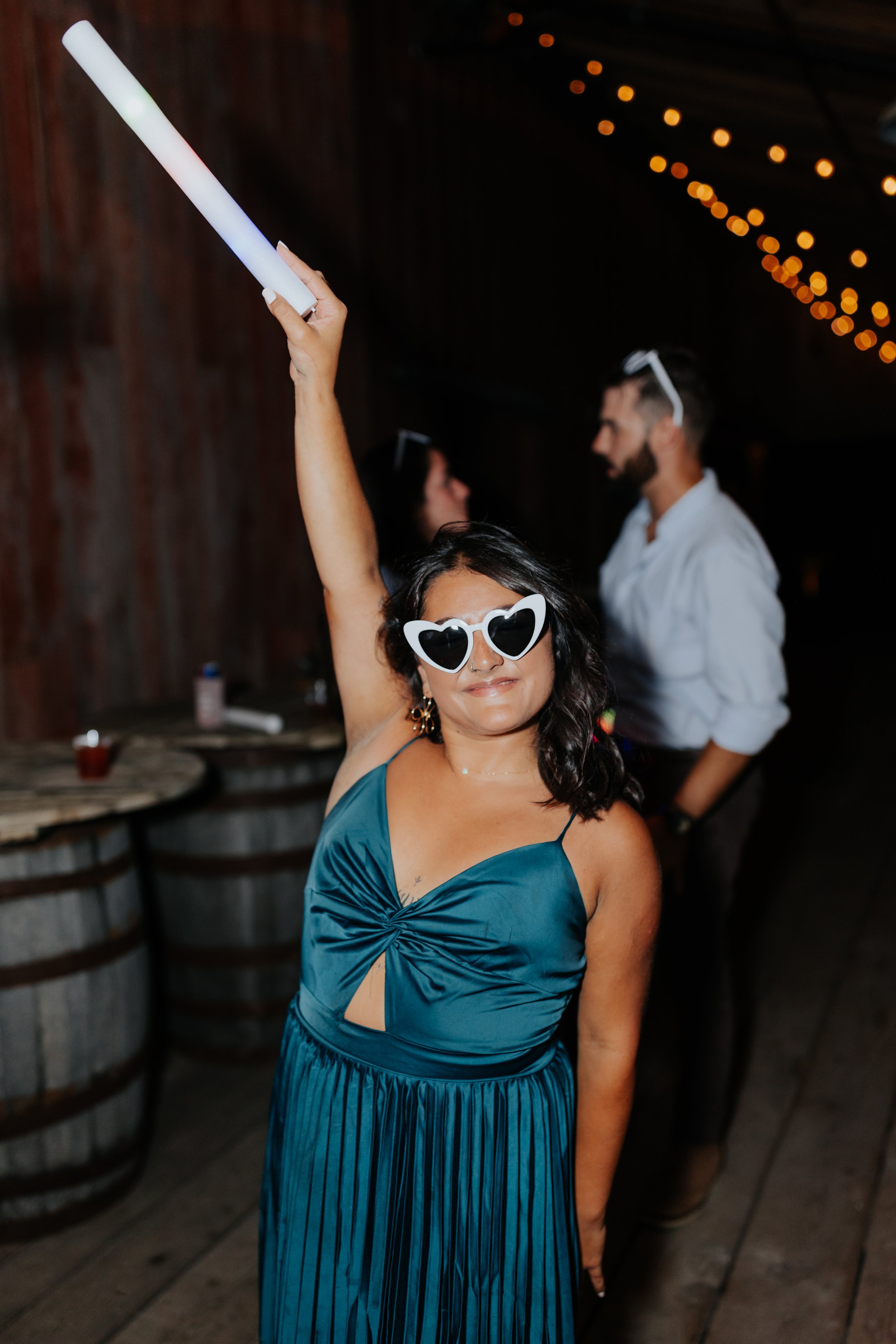 A woman wearing heart-shaped sunglasses and a teal pleated dress standing with her arm raised, holding a glow stick at a party. In the background, there are two people talking, and the setting appears to be a rustic venue with string lights.