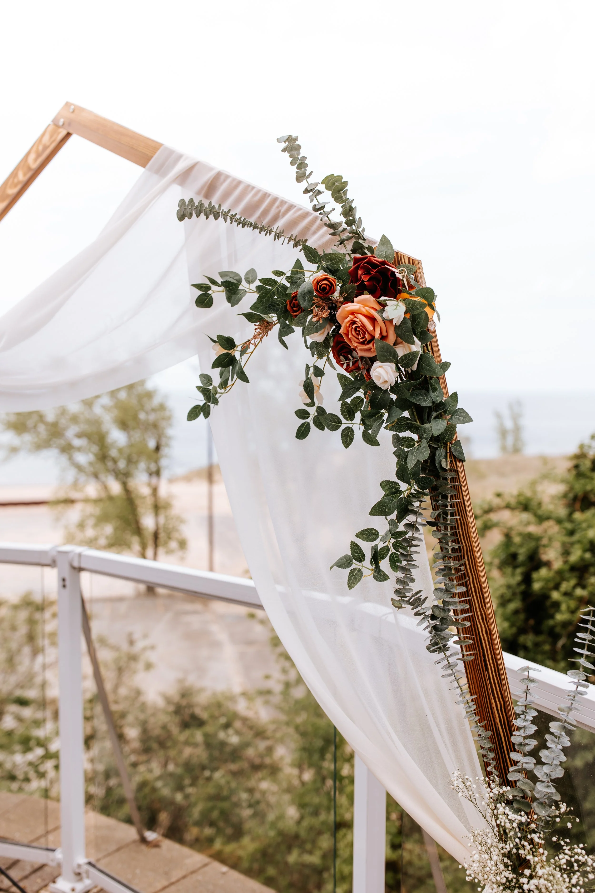 Wedding arch decorated with white sheer fabric and a bouquet of roses and eucalyptus leaves, set outdoors with trees and a cloudy sky in the background.