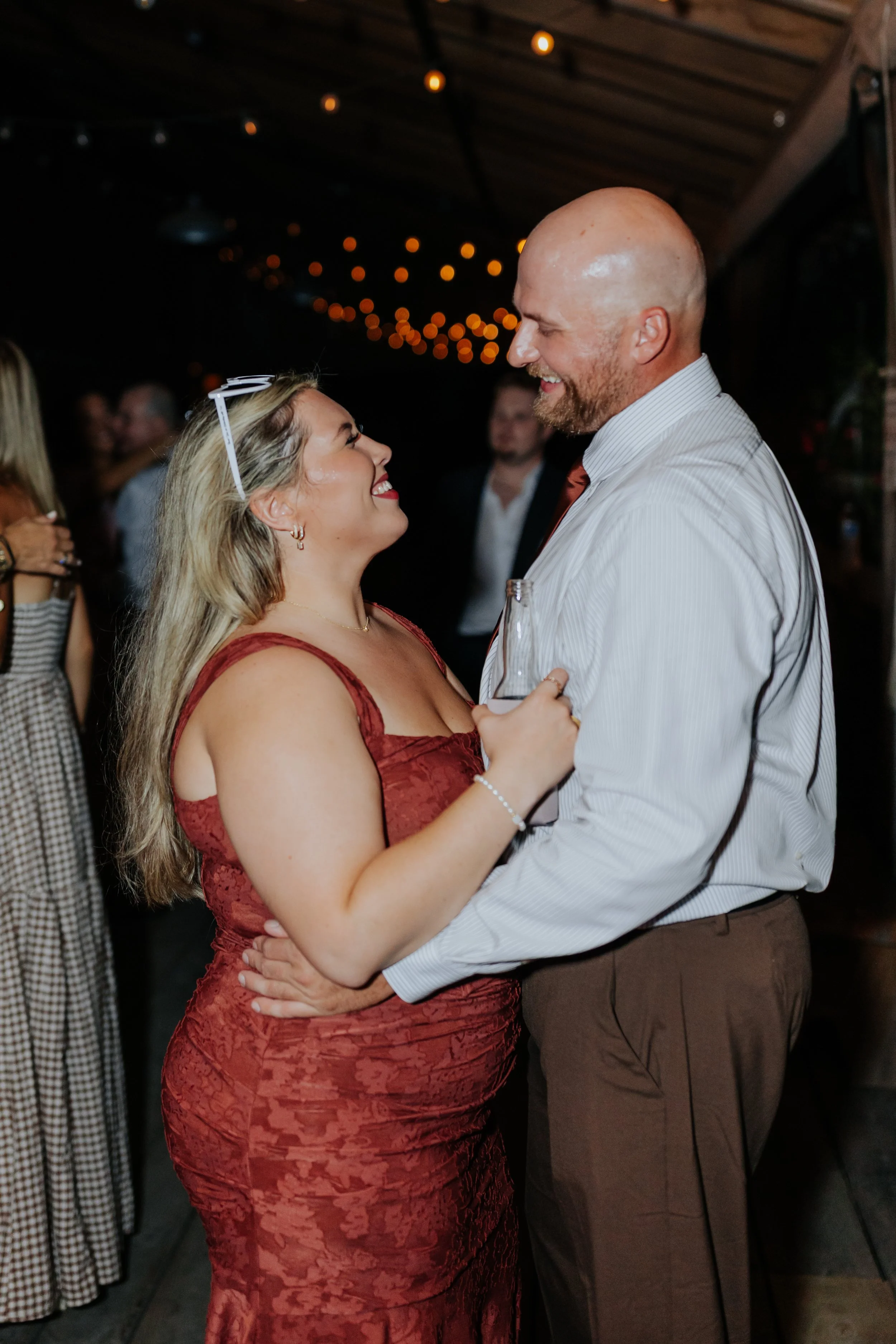 A man and woman sharing a dance at a party or wedding reception, with string lights overhead and other guests in the background.
