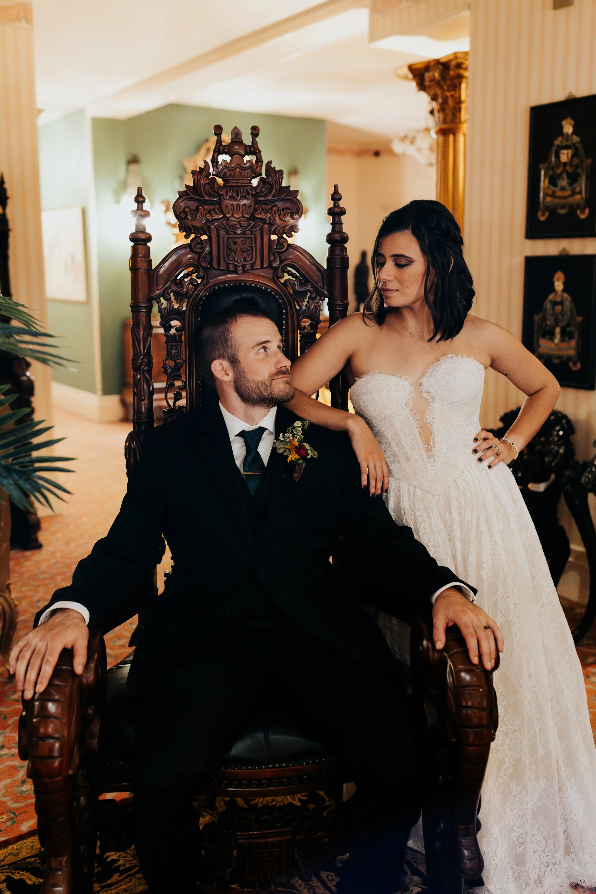 A bride and groom pose in a regal setting, with the groom sitting on an ornate wooden throne and the bride standing beside him, gazing at him.