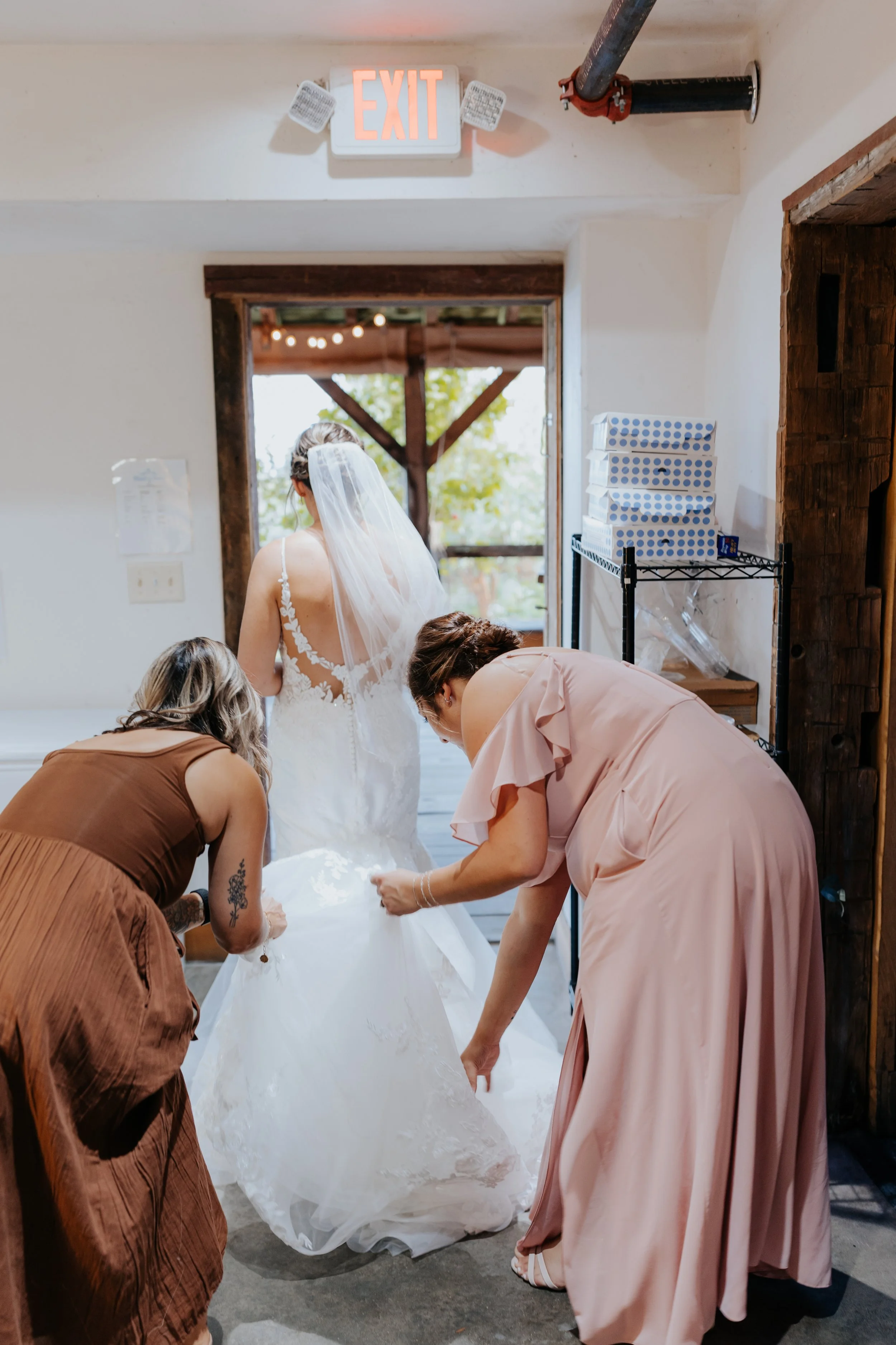 Three women helping a bride with her wedding dress inside a rustic room with an open door leading outside. There is an illuminated exit sign overhead.
