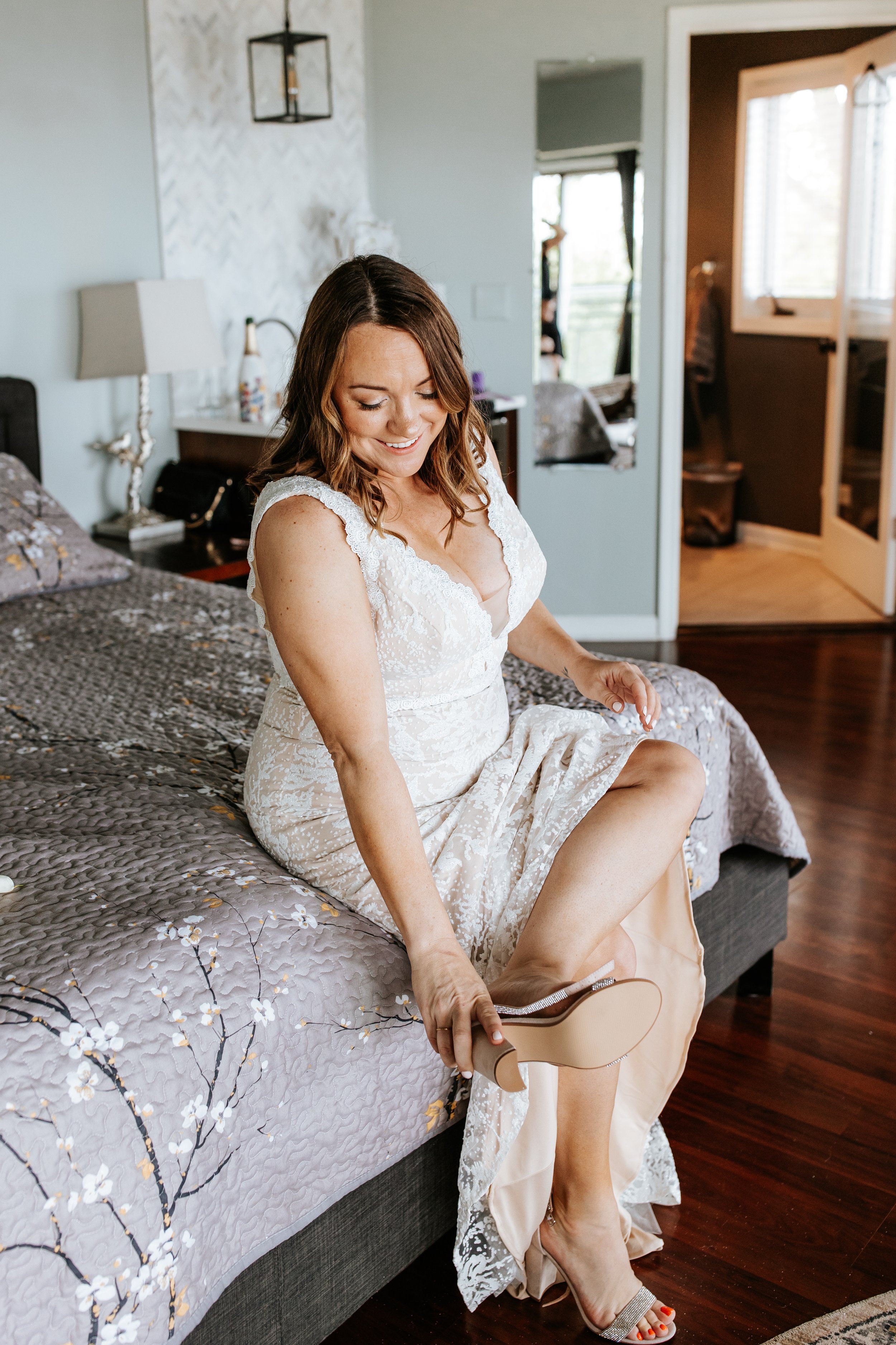 A woman in a white lace dress sitting on a bed, putting on a high heel shoe, smiling.