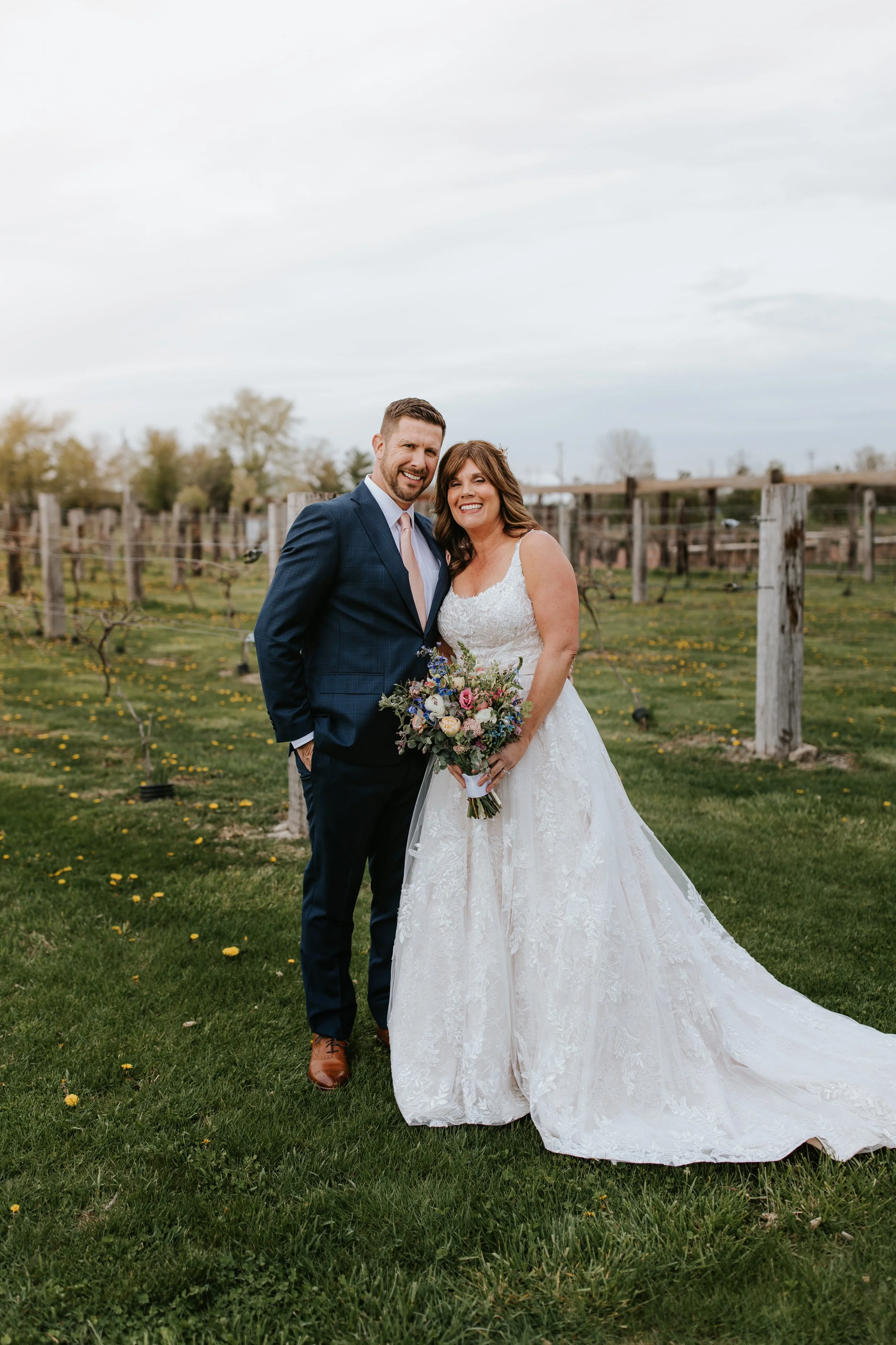 A bride and groom standing outdoors in a grassy area with flowering trees and a vineyard in the background, smiling.