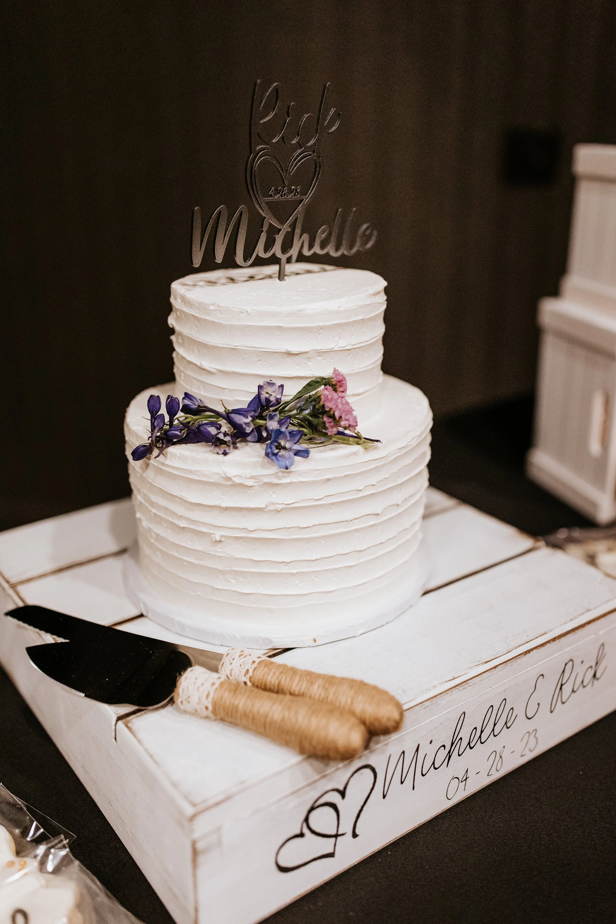 Two-tiered white wedding cake with purple and pink flowers, a 'Love Michelle' cake topper, placed on a white wooden tray with a cake knife and server, with the names Michelle and Rick and the date 04-28-23 written on the tray.