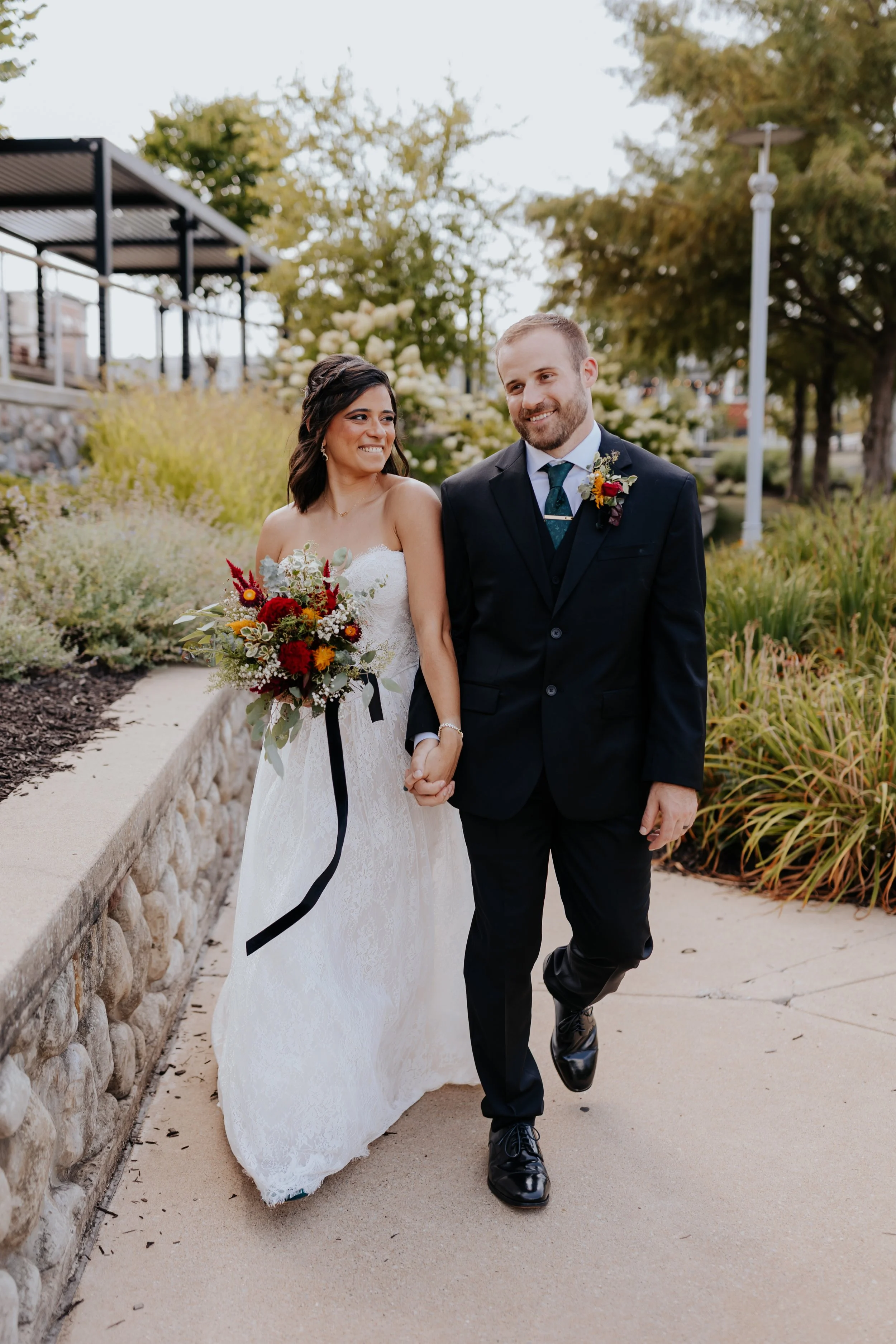 A newlywed couple walking outdoors, holding hands, with a woman in a white wedding dress holding a bouquet of flowers, and a man in a black suit. The setting includes greenery, trees, and a stone wall.