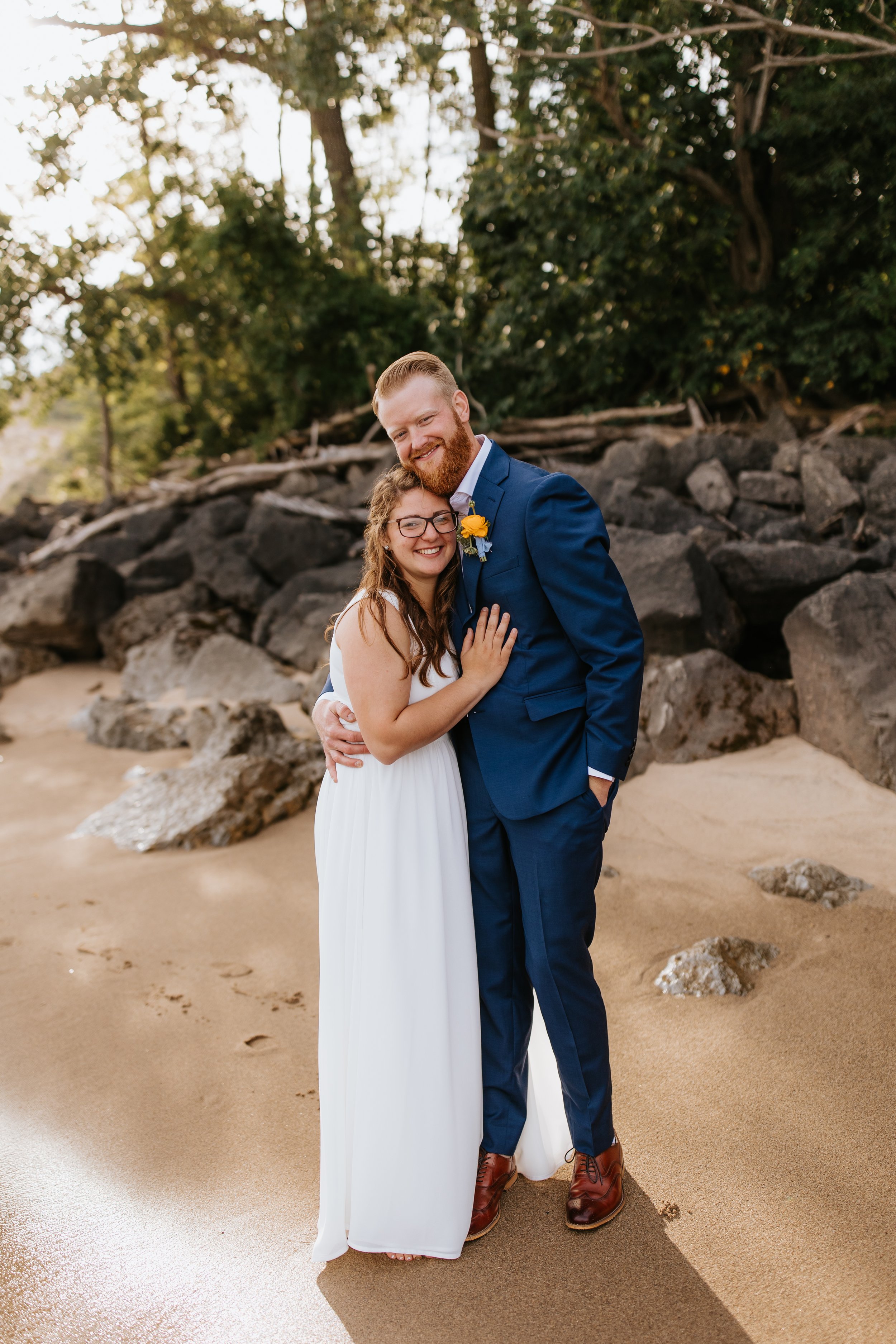A smiling couple embracing on the beach, the woman in a white dress and the man in a blue suit, with rocks and trees in the background.