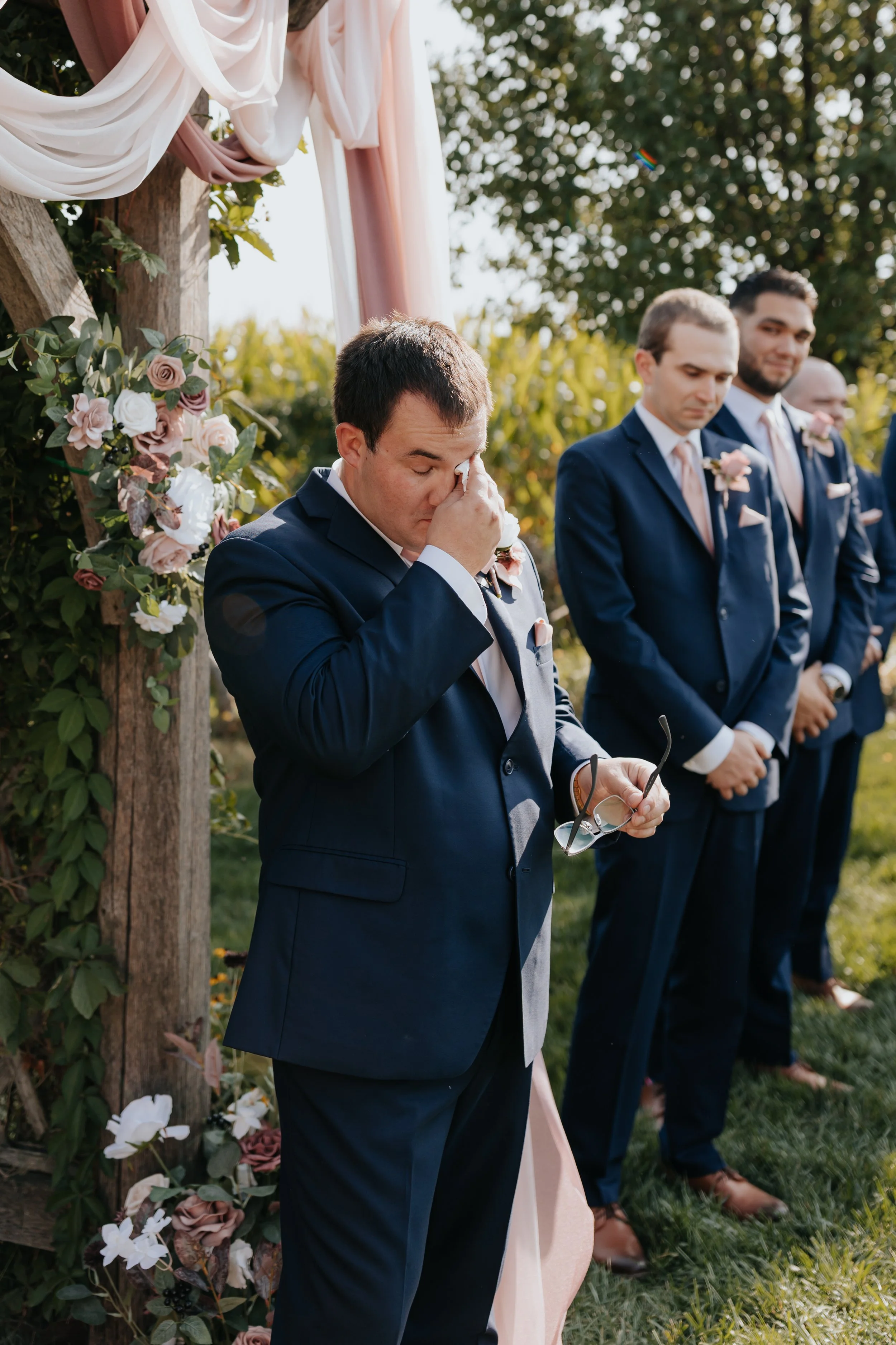 A man in a navy suit is wiping his eyes during an outdoor wedding ceremony, standing in front of a floral arch with greenery and pink and white flowers. Other men in navy suits with pink ties stand behind him, some with bowed heads.