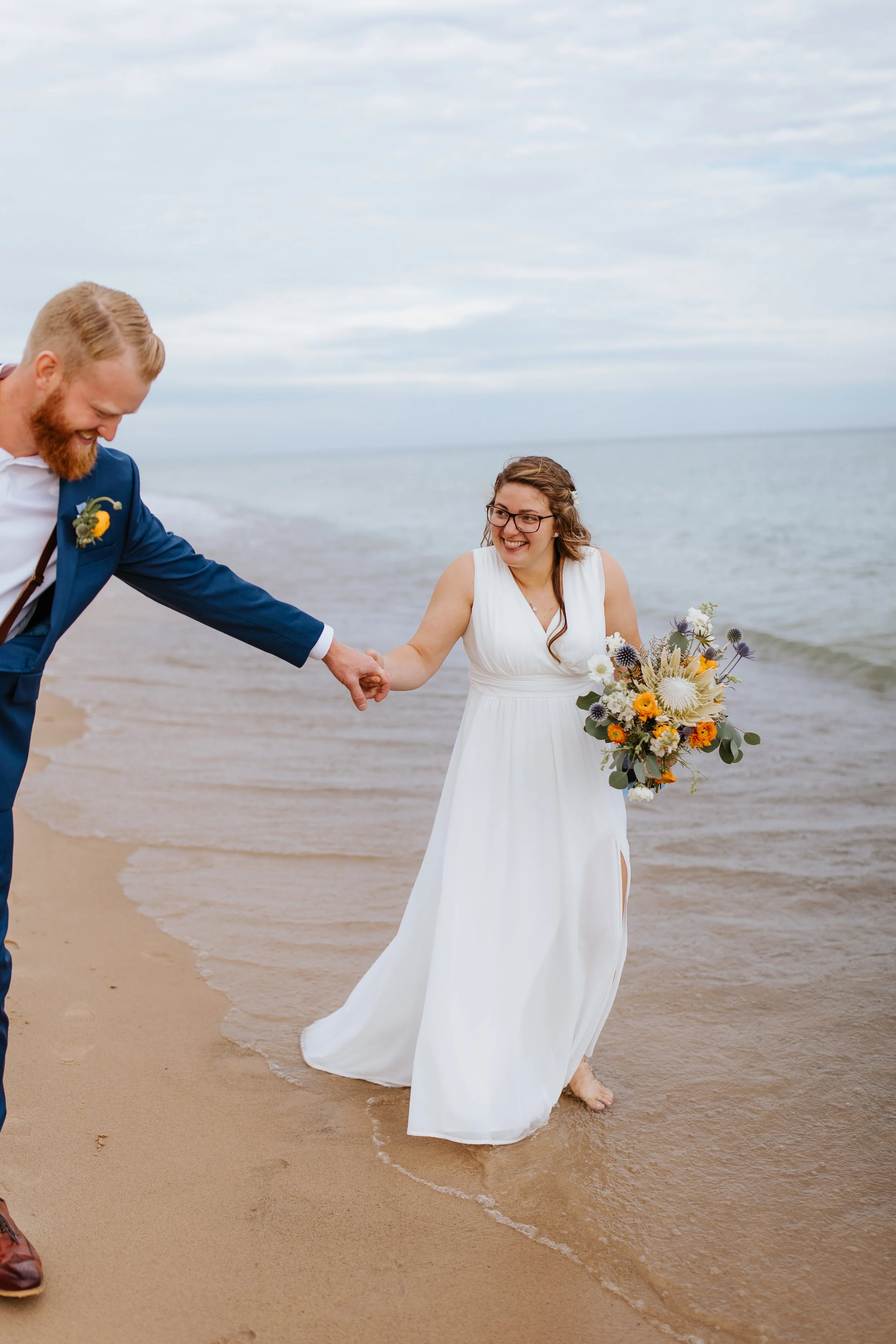 A bride and groom holding hands and walking barefoot on the beach, smiling and enjoying their wedding on the shoreline.