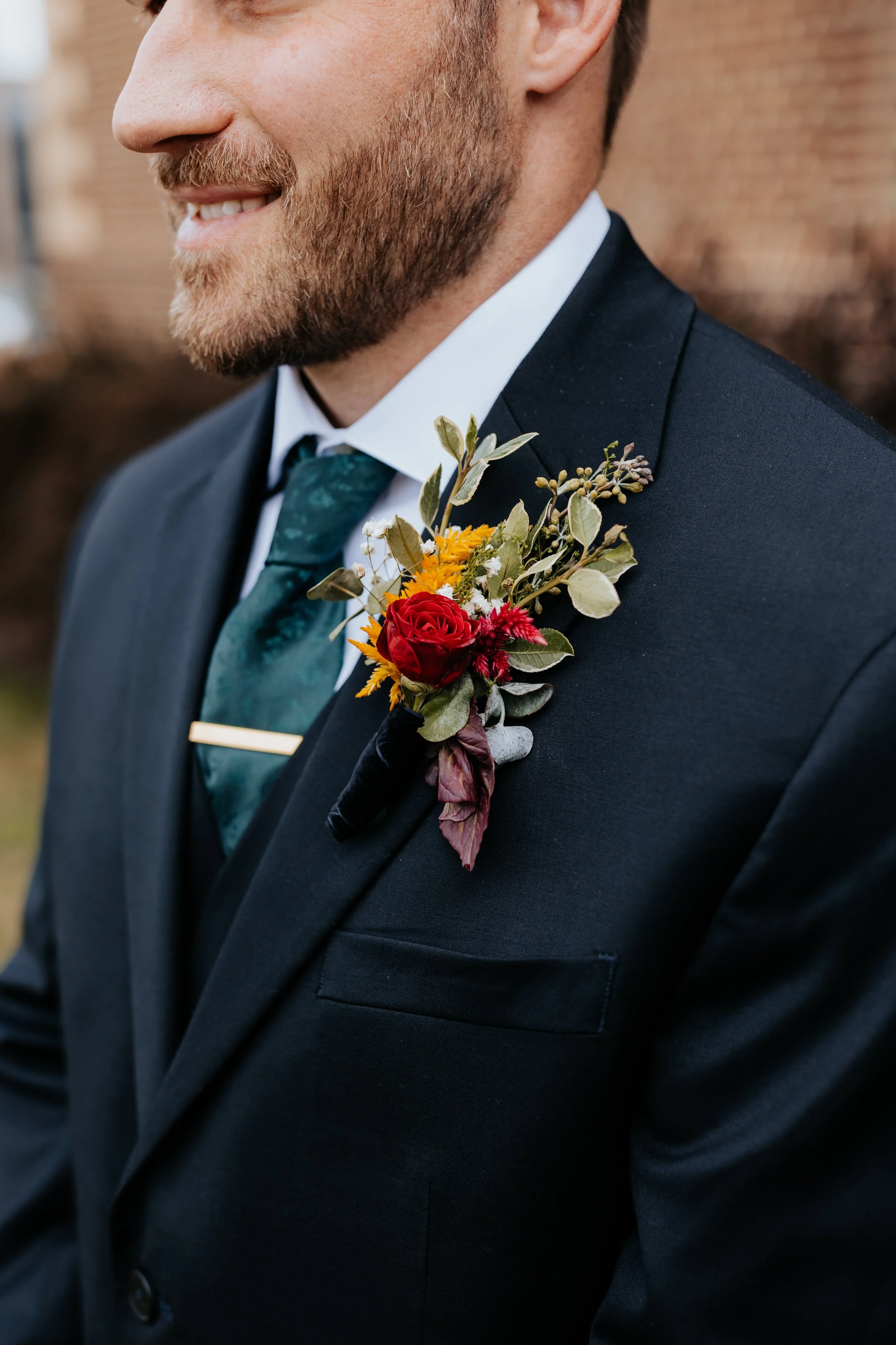 Close-up of a man dressed in formal attire wearing a dark suit, white shirt, and teal tie, with a colorful boutonniere on his lapel featuring red, yellow, and purple flowers along with greenery, outdoors during daytime.