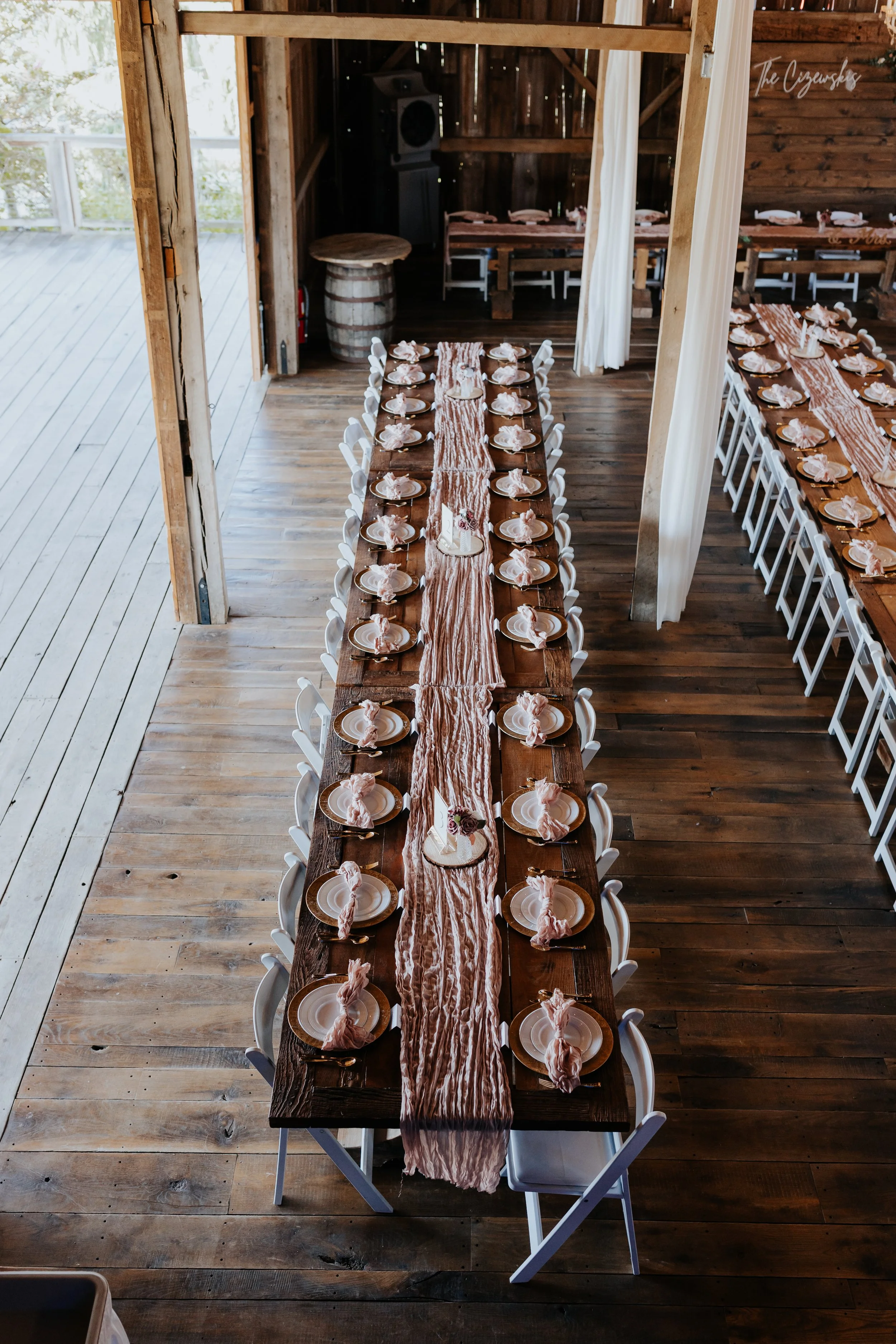 Long wooden dining table set for a celebration with plates, napkins, and small decorations, inside a rustic wooden building with a wooden floor and outdoor view.