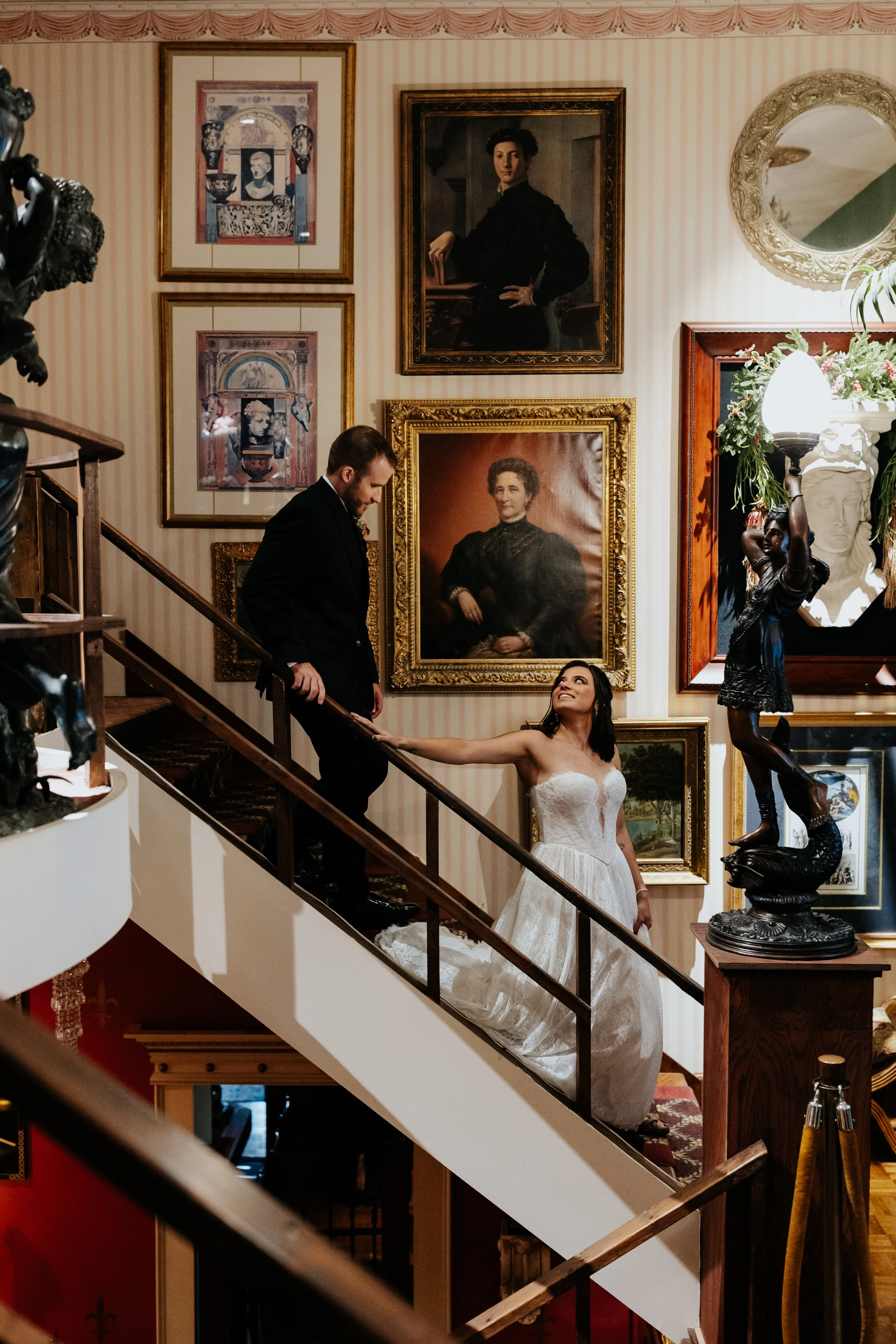 A bride and groom on a staircase inside an art gallery, surrounded by framed paintings and sculptures.