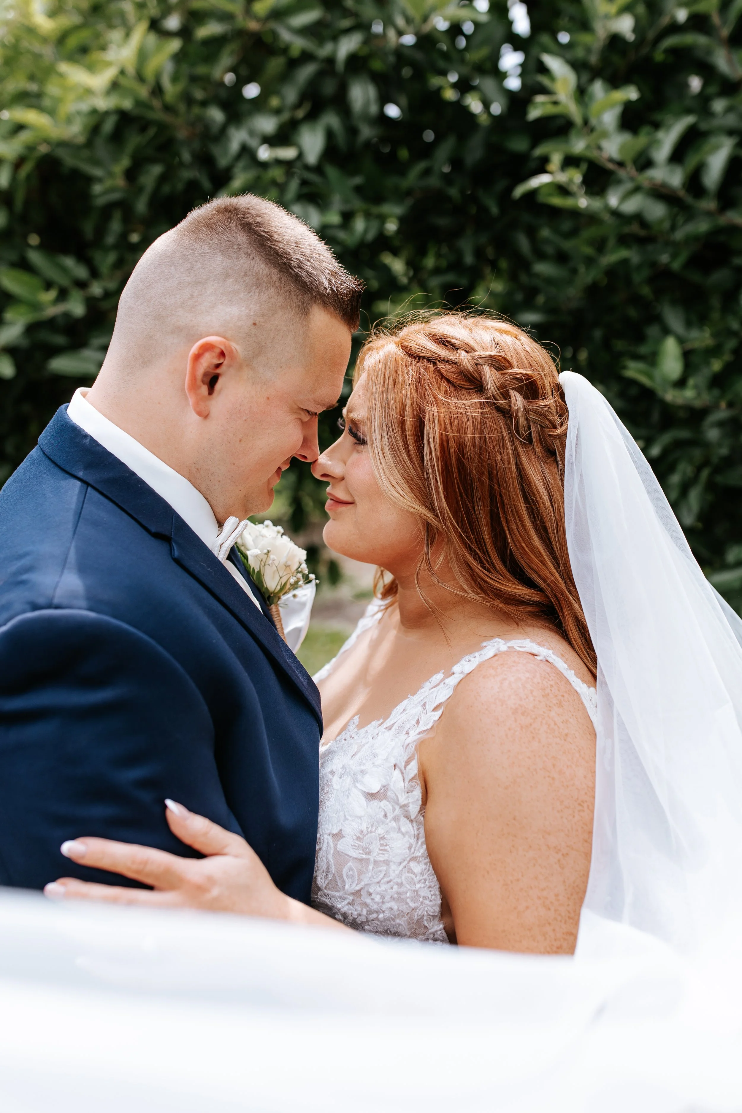 Close-up of a bride and groom touching foreheads, outdoors, with green foliage in the background.