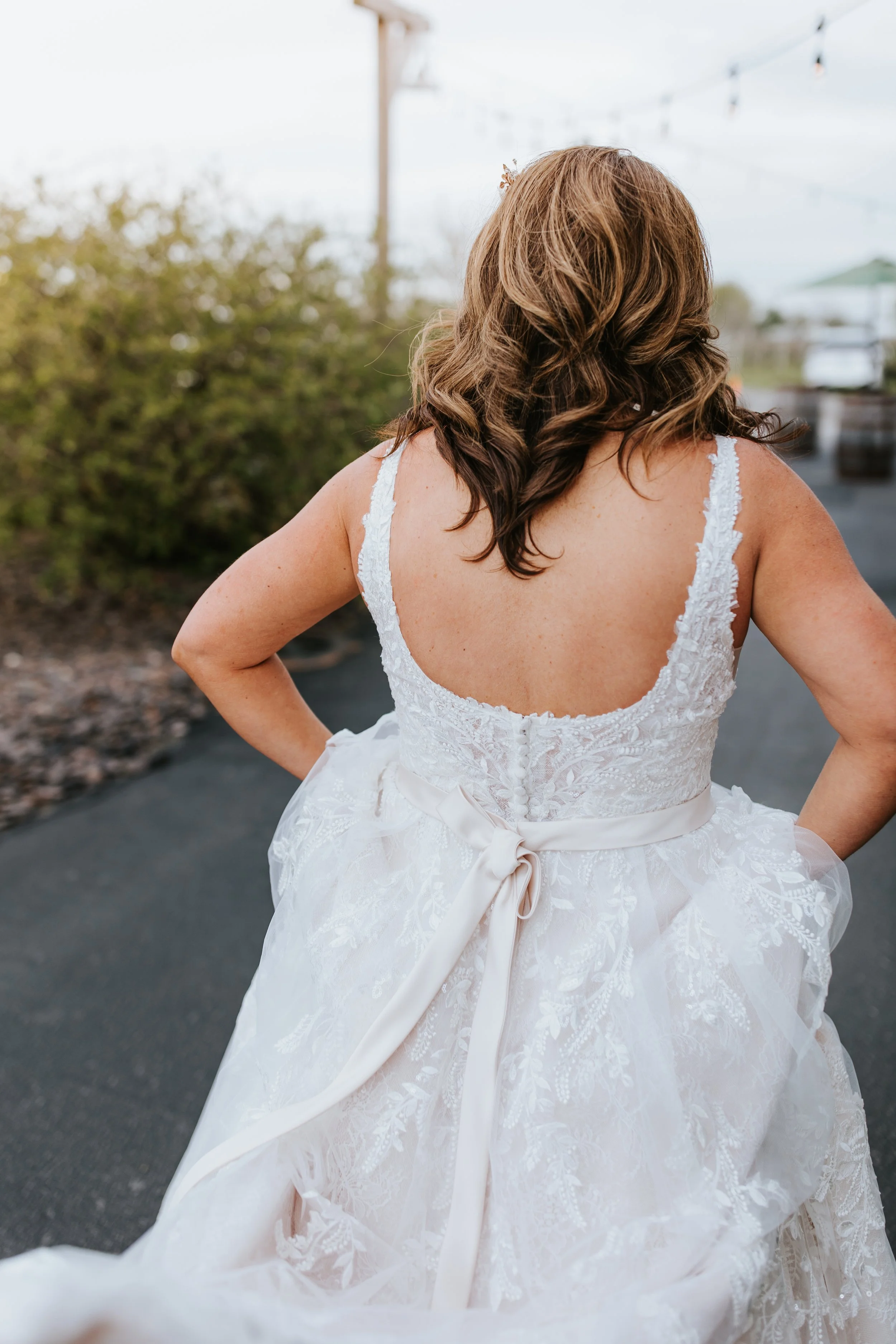 Back view of a woman in a white lace wedding dress with a satin ribbon tied in a bow, standing outdoors.