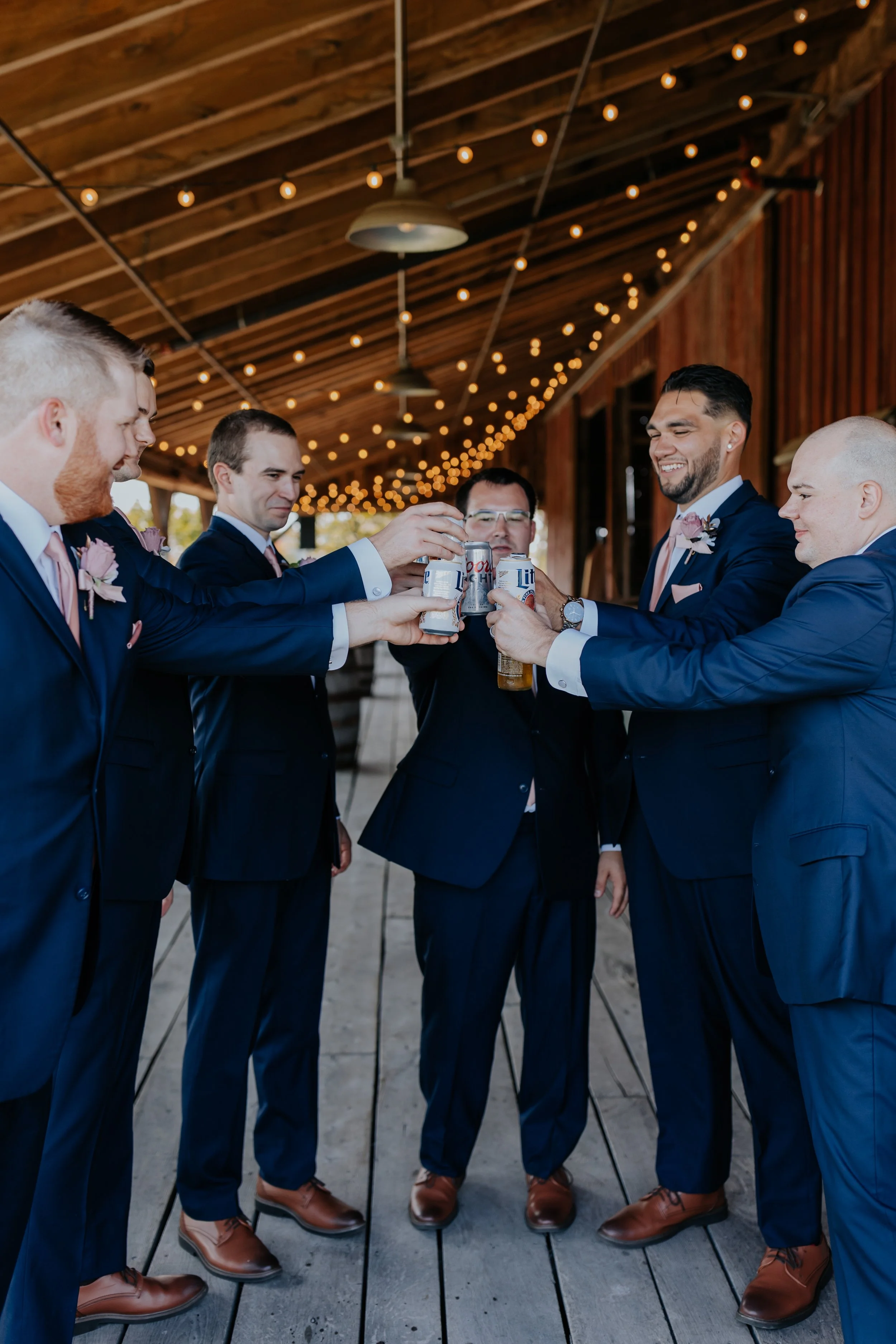Groom and groomsmen toast with beer at wedding reception under string lights in rustic venue.
