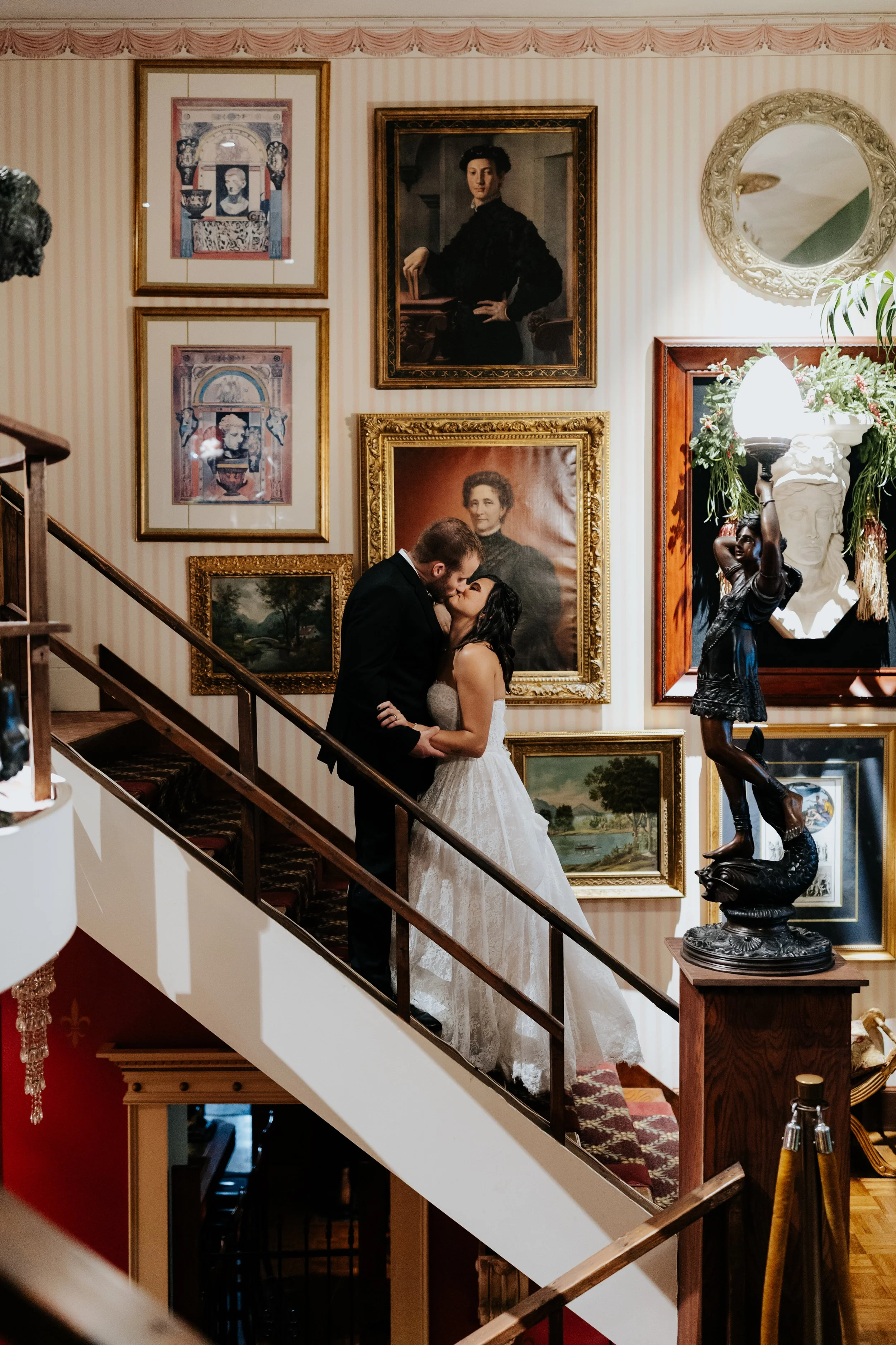 A couple dressed in wedding attire sharing a kiss on a staircase surrounded by framed portraits and paintings on the wall.