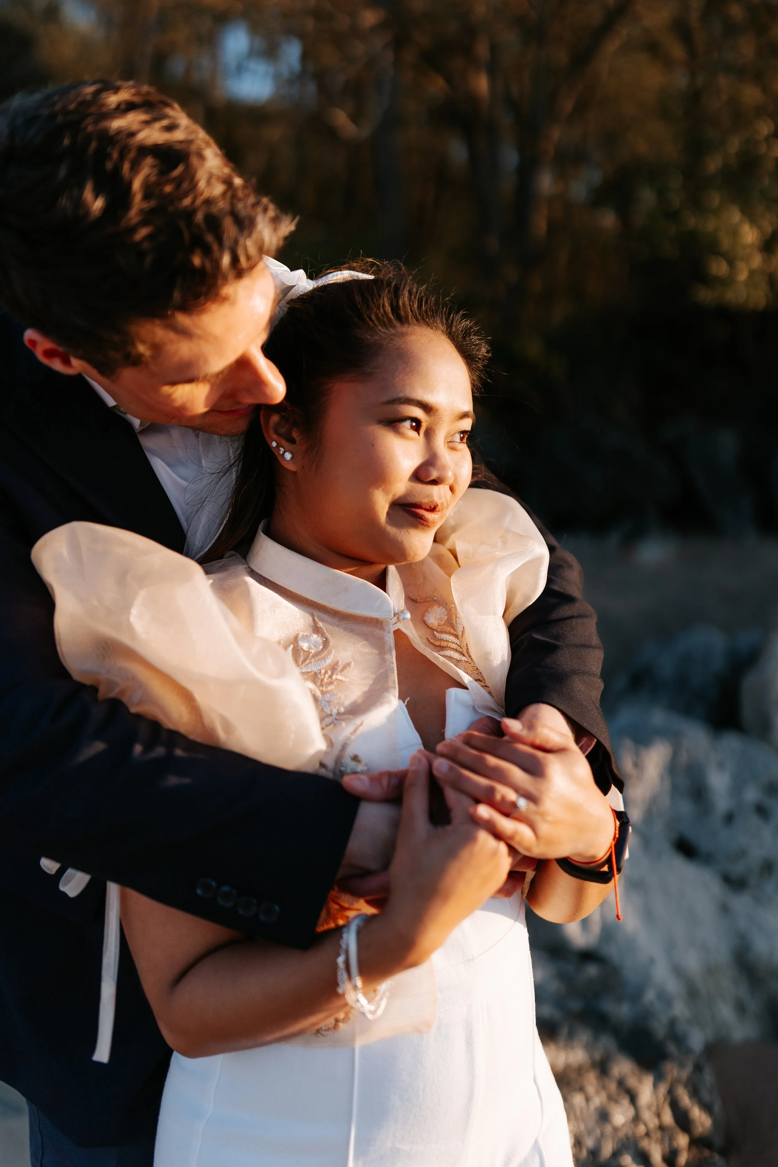 A couple embracing outdoors during sunset, with trees in the background, as the man kisses the woman on the side of her head.
