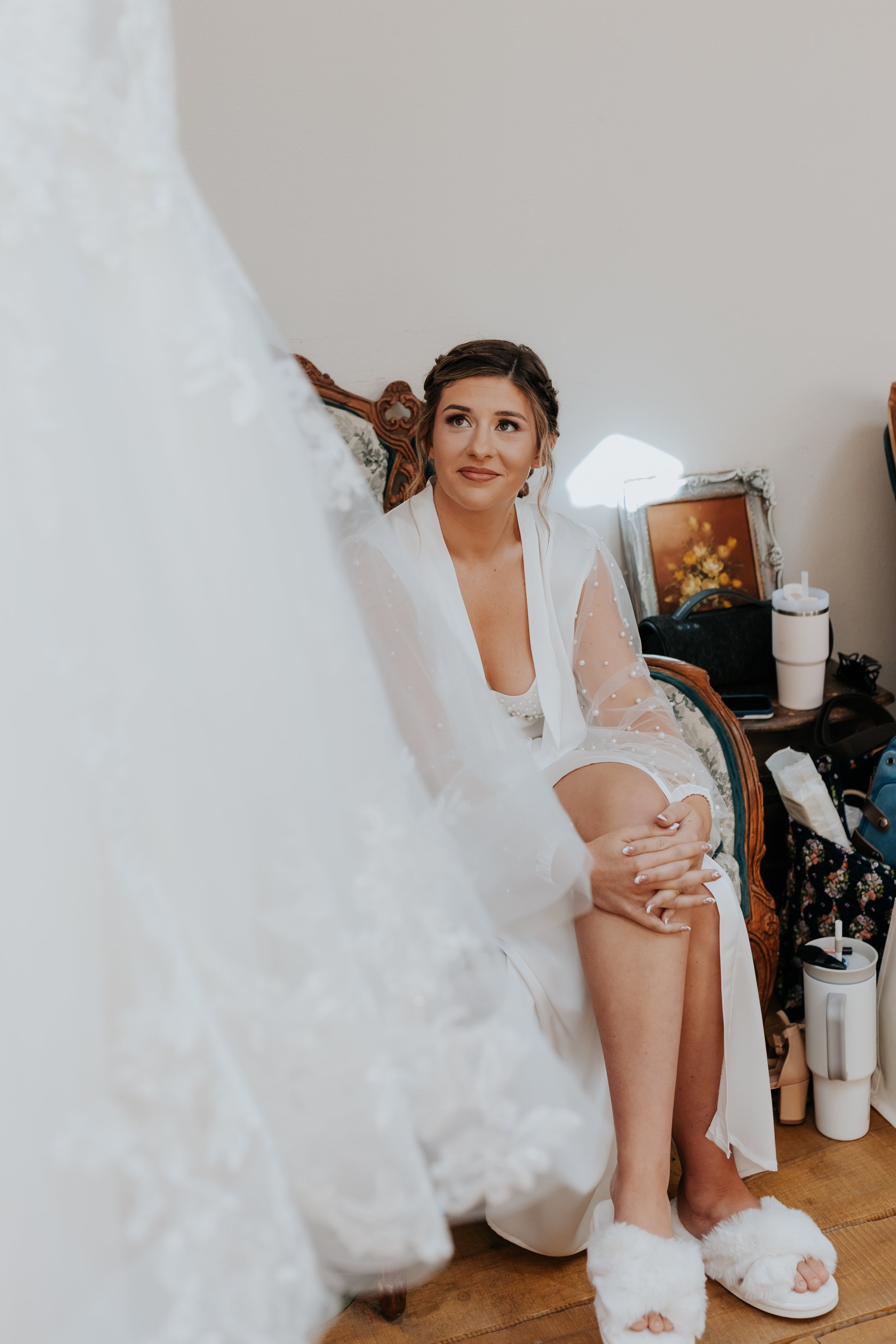 A woman sitting on a vintage wooden chair, wearing a white robe, looking up with a gentle smile, surrounded by personal items and a framed painting on a side table.