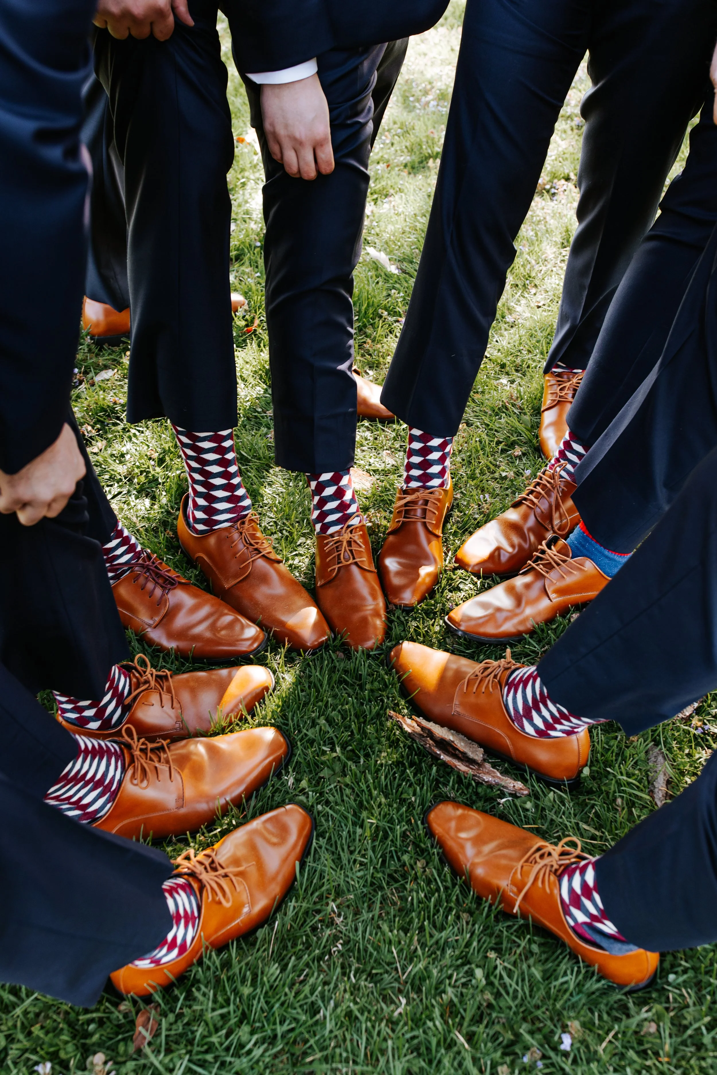 A group of men dressed in navy suits and brown dress shoes standing in a circle outdoors, with their shoes pointed toward the center, on grass. They are wearing colorful patterned socks.