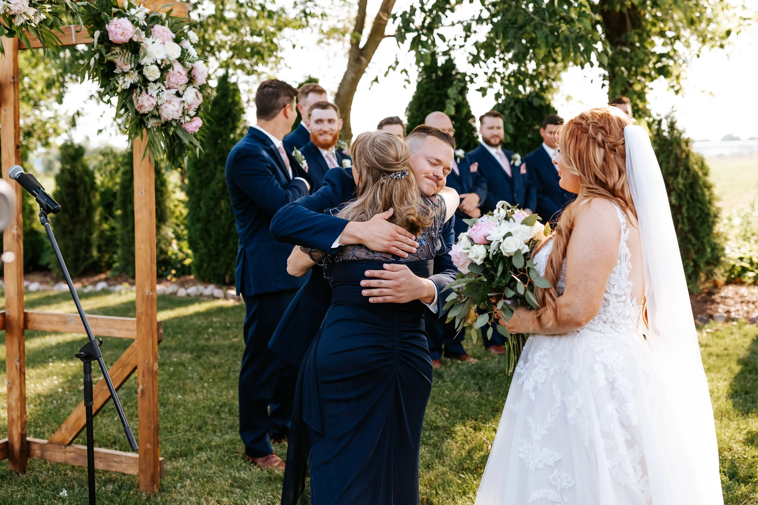 A bride in a white wedding dress holding a bouquet of flowers at an outdoor wedding ceremony, with a groom and others in formal attire standing behind her; guests are embracing and smiling under a decorated wooden arch, with trees and greenery in the