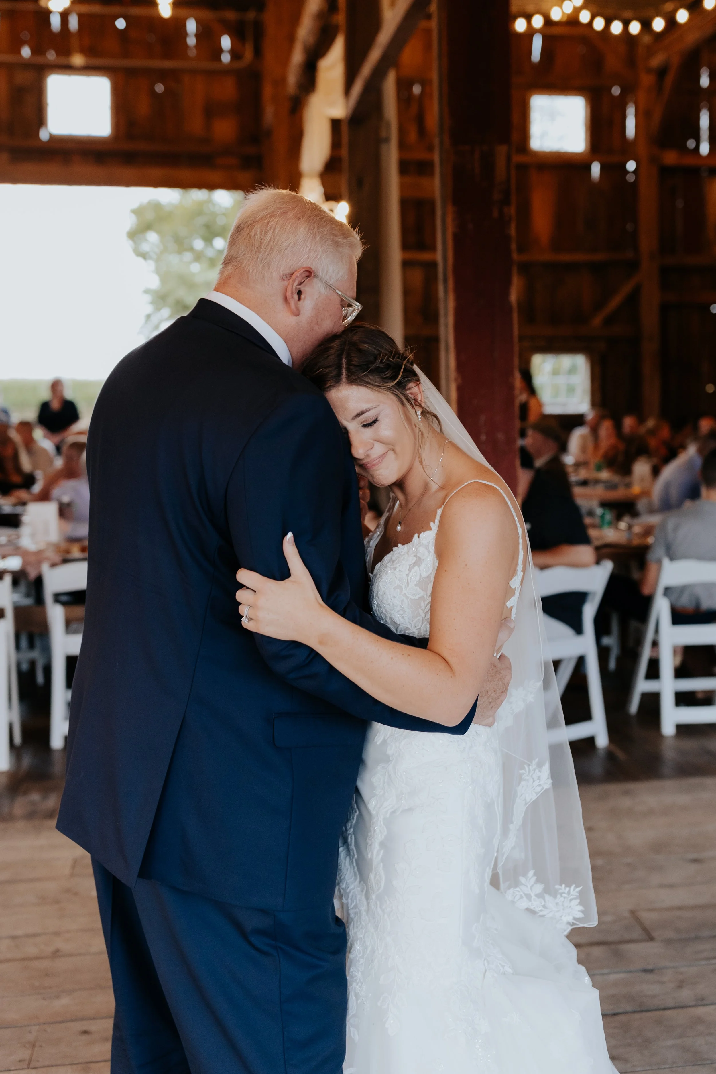 A bride and groom share a tender moment during their wedding inside a rustic barn with wooden walls, seated guests, and natural light.