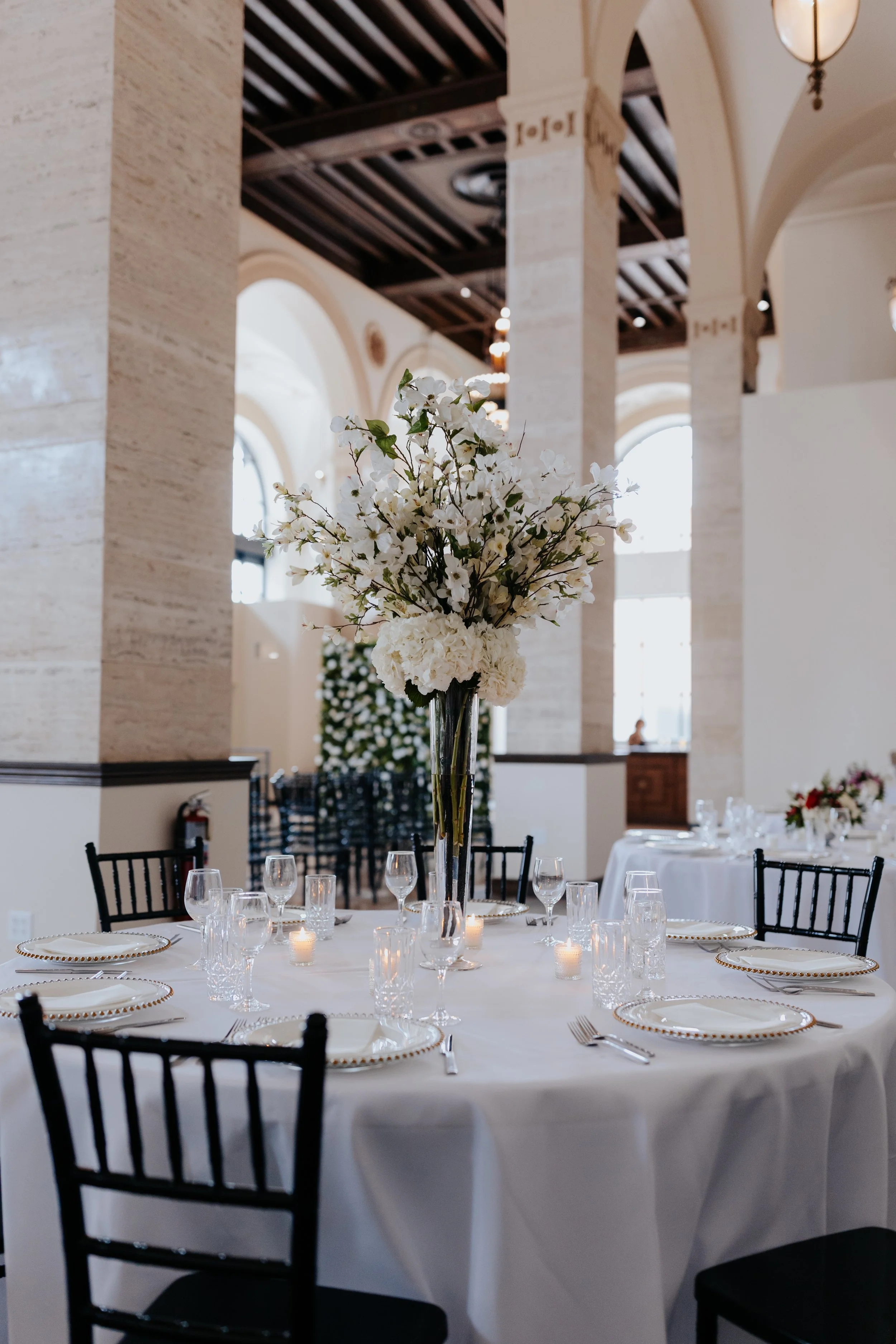 Elegant indoor event space with a round table decorated with a tall floral centerpiece of white flowers, surrounded by black chairs, set for a formal occasion with plates, glasses, and candles.