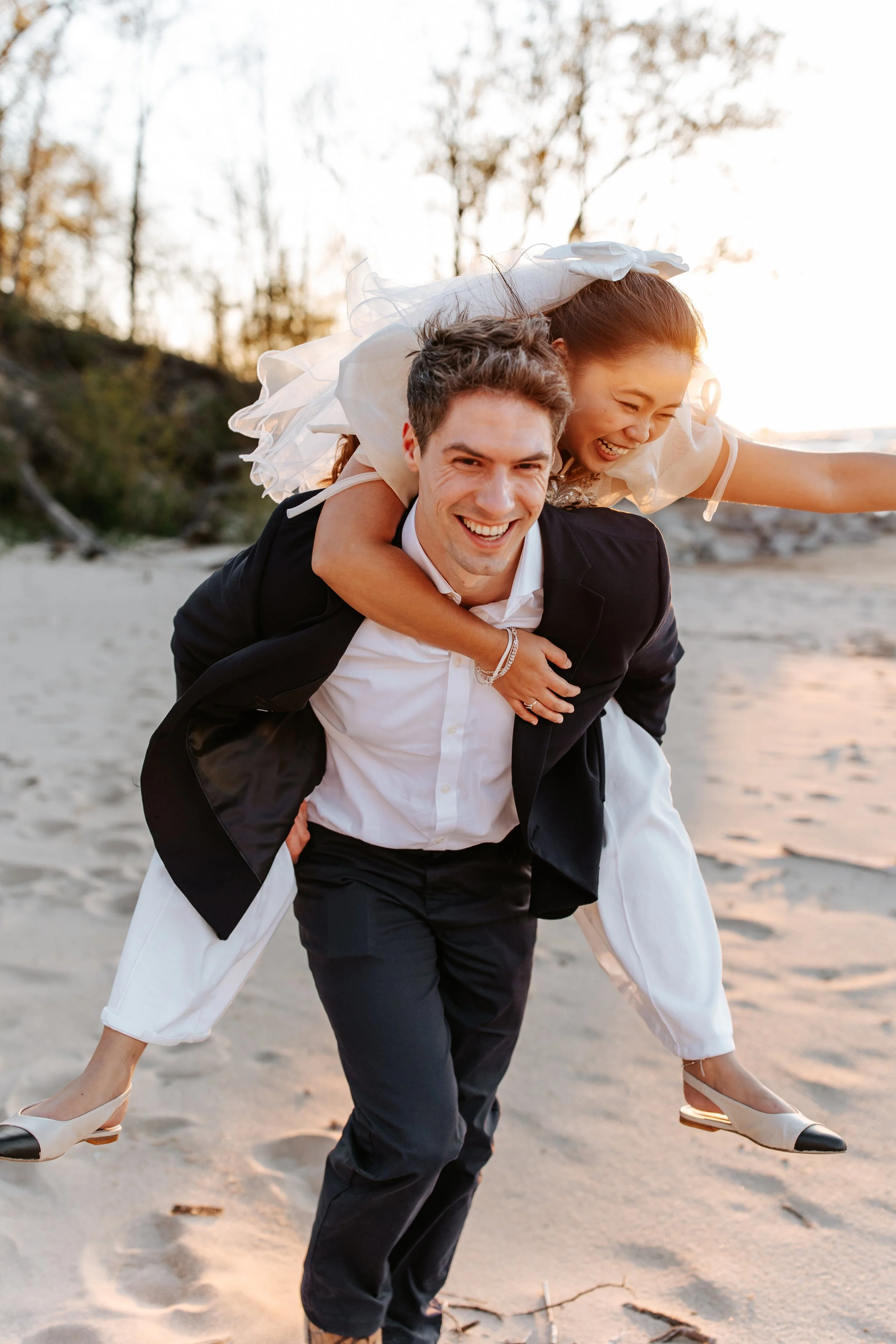A man giving a piggyback ride to a woman on a beach. Both are smiling and appear happy, with trees and water in the background at sunset.