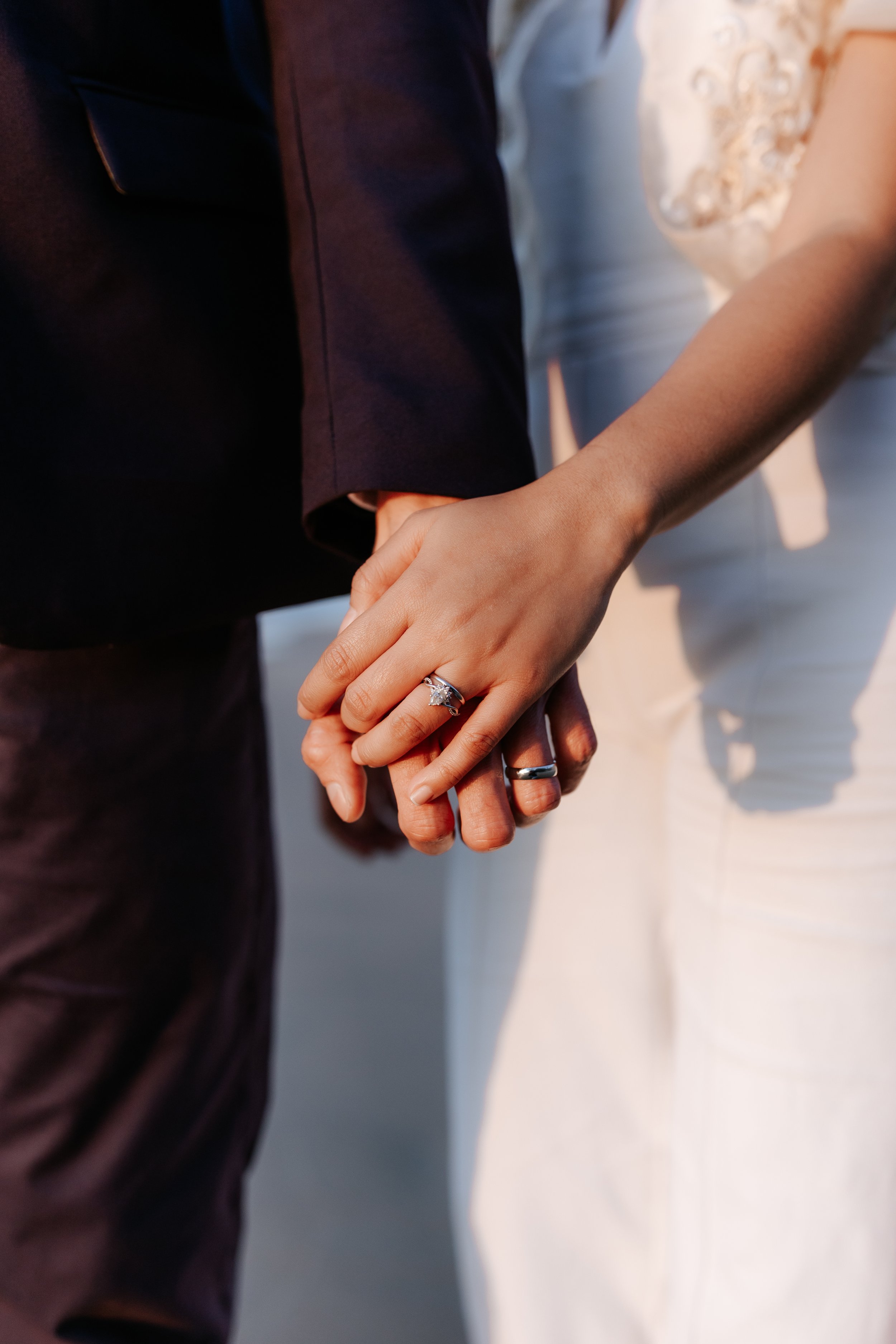 Close-up of a bride and groom holding hands, showing wedding rings, with the bride in a white dress and the groom in a dark suit.