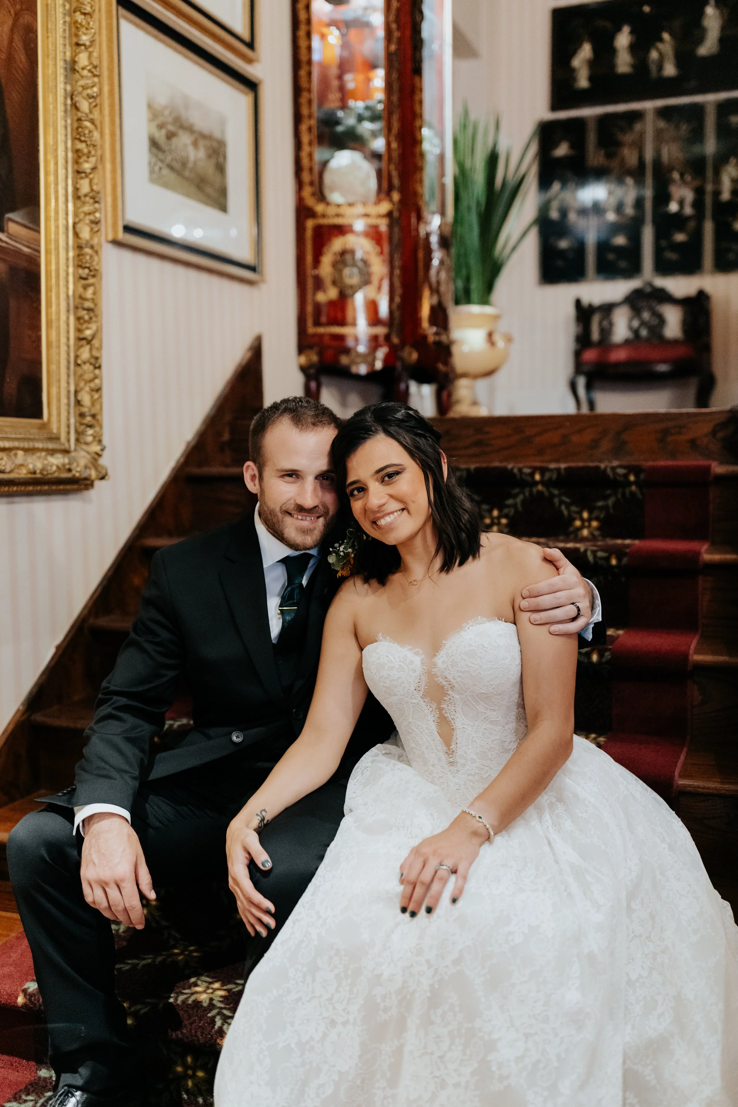 A smiling bride and groom sitting close together on a staircase, with the bride in a white lace wedding dress and the groom in a black suit, in a decorated indoor setting.