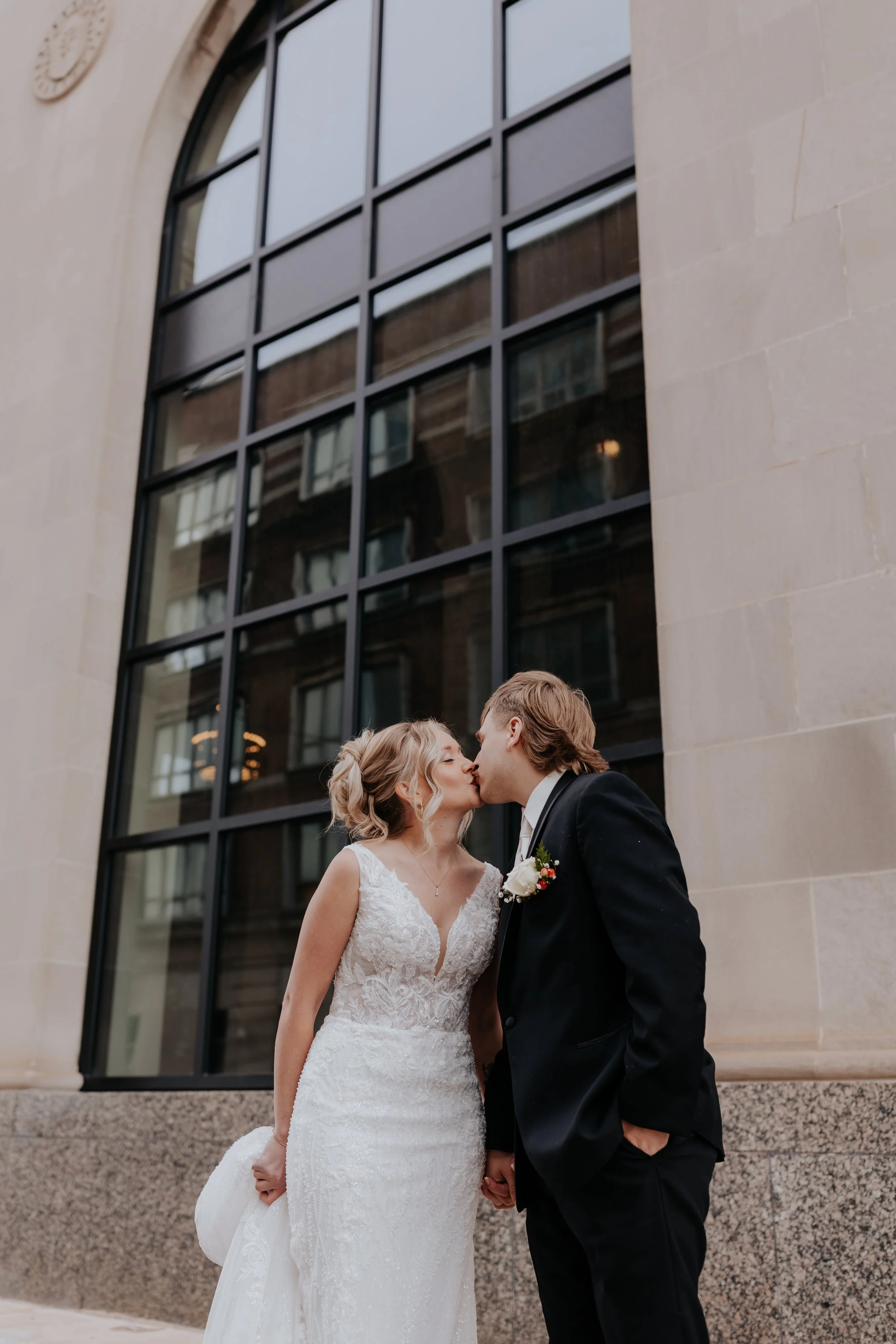 A bride and groom kissing outside a building with large black-framed windows.