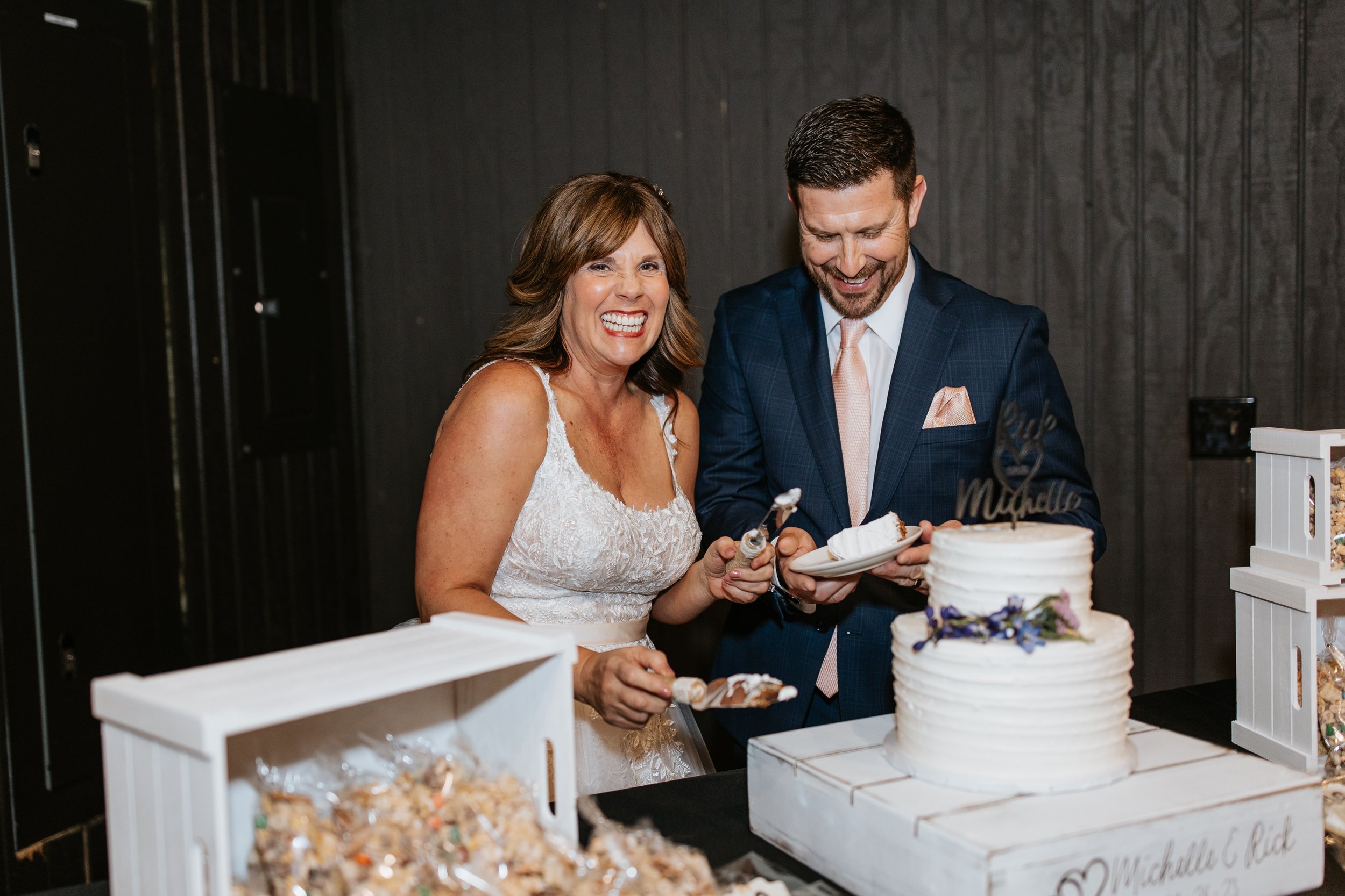 A woman in a white wedding dress and a man in a dark suit with a pink tie are smiling and cutting a wedding cake together at their wedding reception. The wedding cake is white with purple flowers on it. They are surrounded by wedding treats and decor