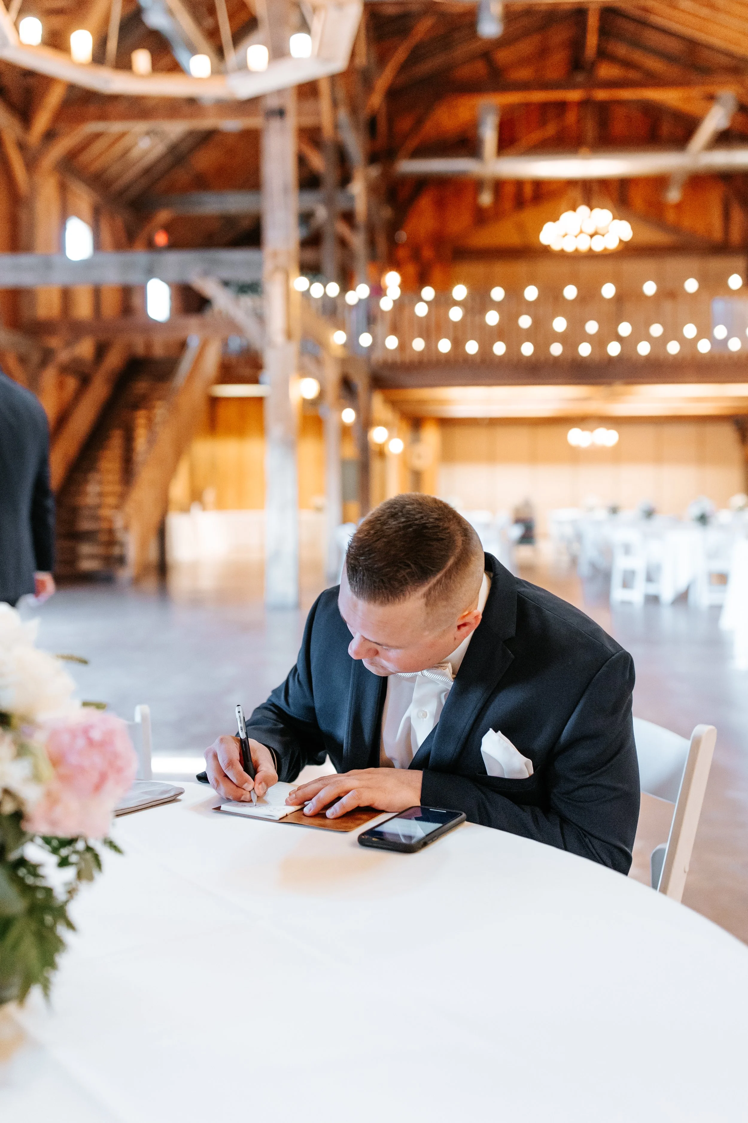 A man in a black suit signing a book at a wedding reception in a rustic barn with string lights.