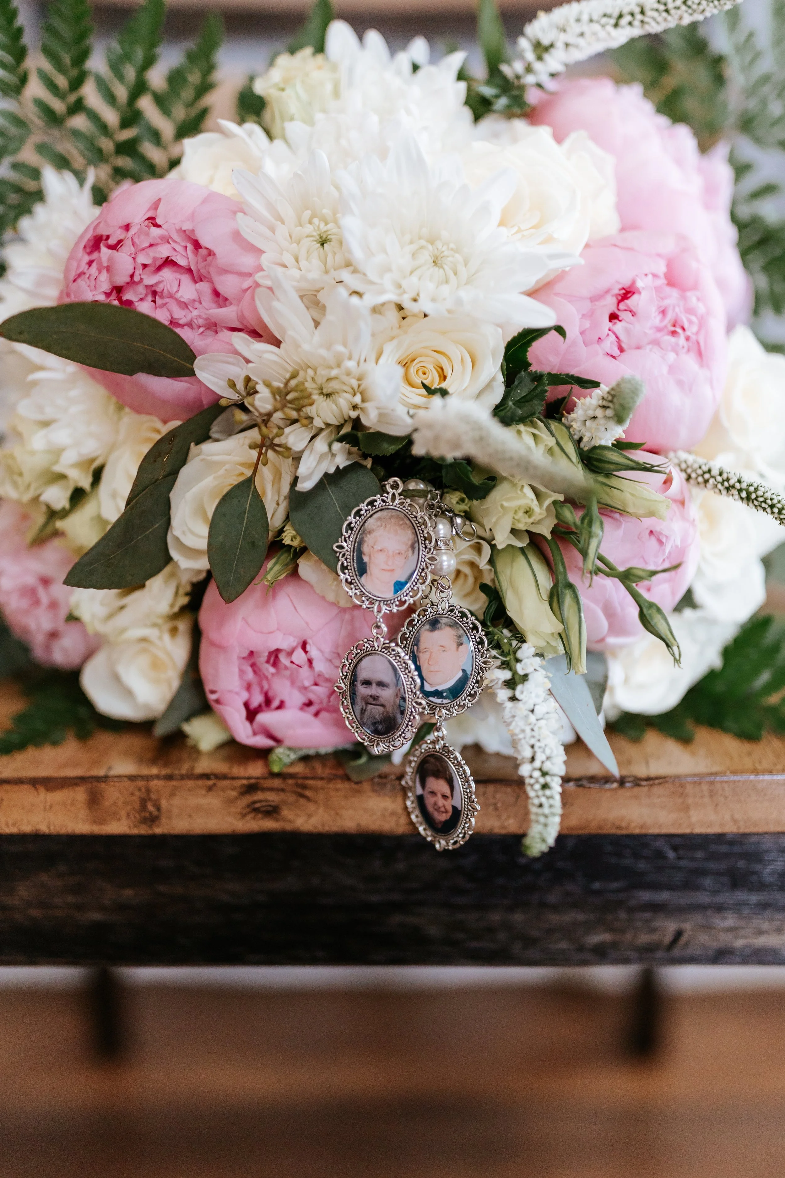 A bouquet of white and pink flowers with green leaves, featuring a hanging picture frame jewelry with four photographs of different women and a man.