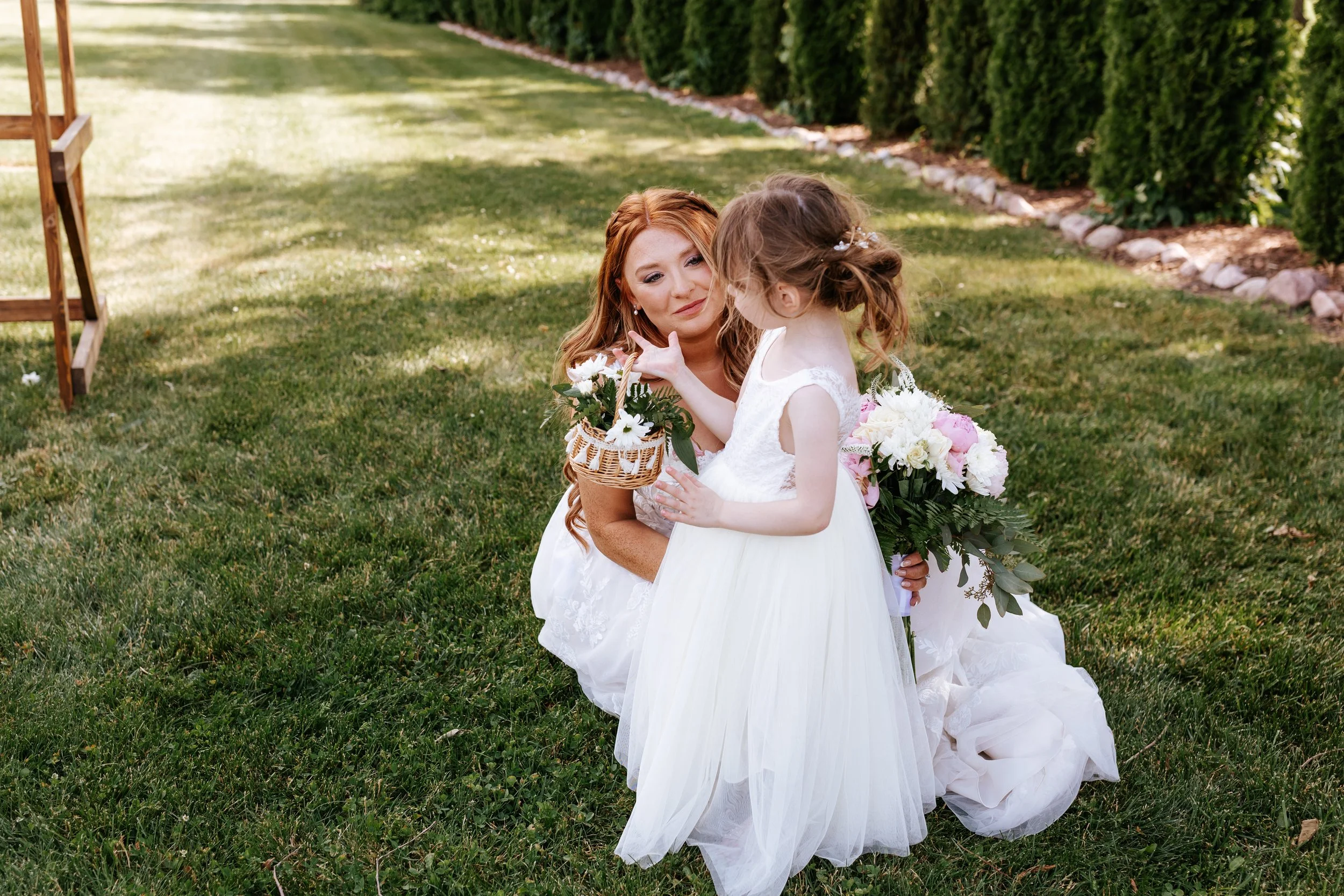 A woman with long red hair in a white dress kneeling on grass, smiling at a young girl in a white dress holding a basket of flowers, during an outdoor wedding or event.