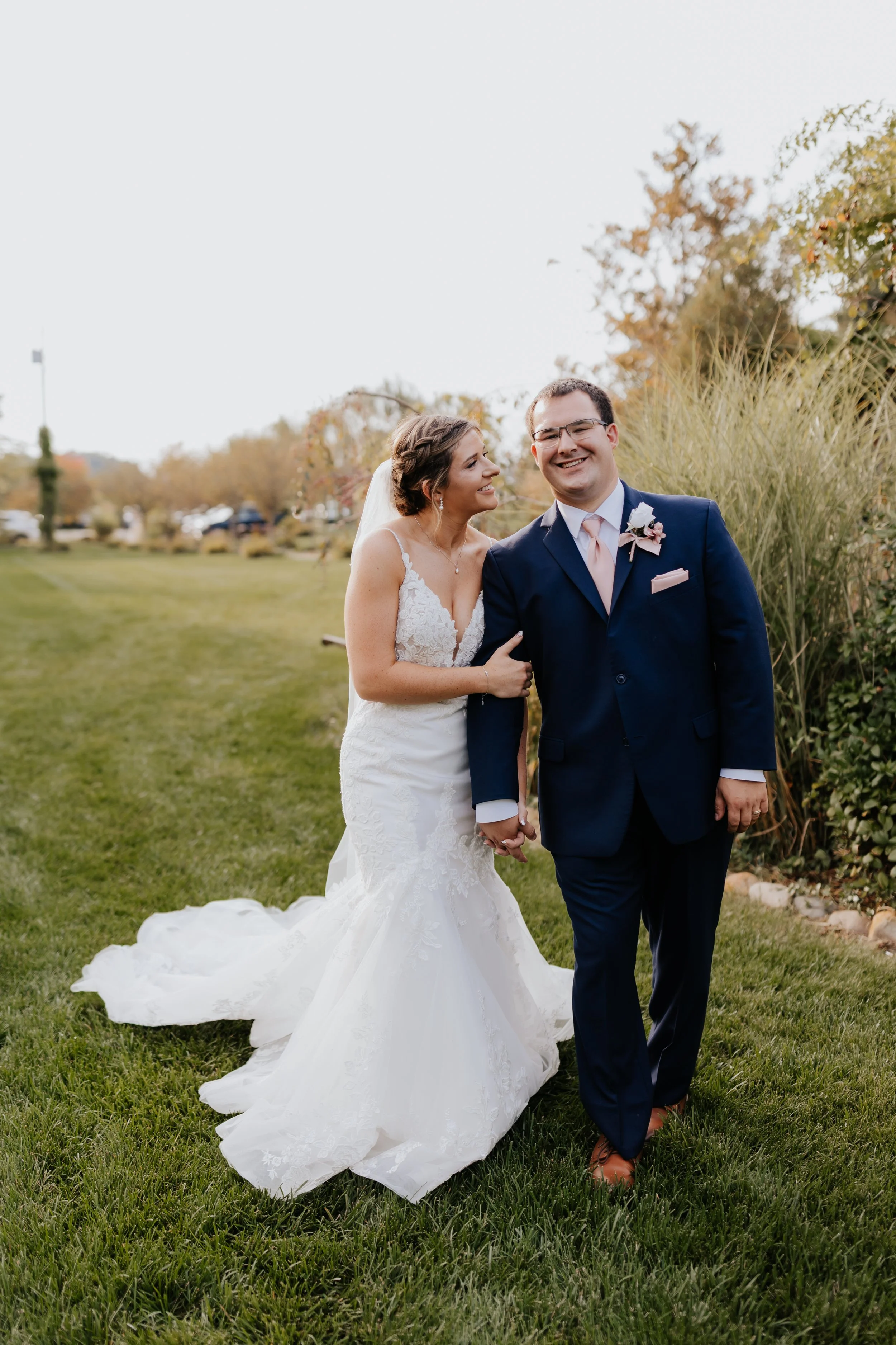 A newlywed couple walking on grass, smiling and holding hands, outdoors with trees and a cloudy sky in the background.