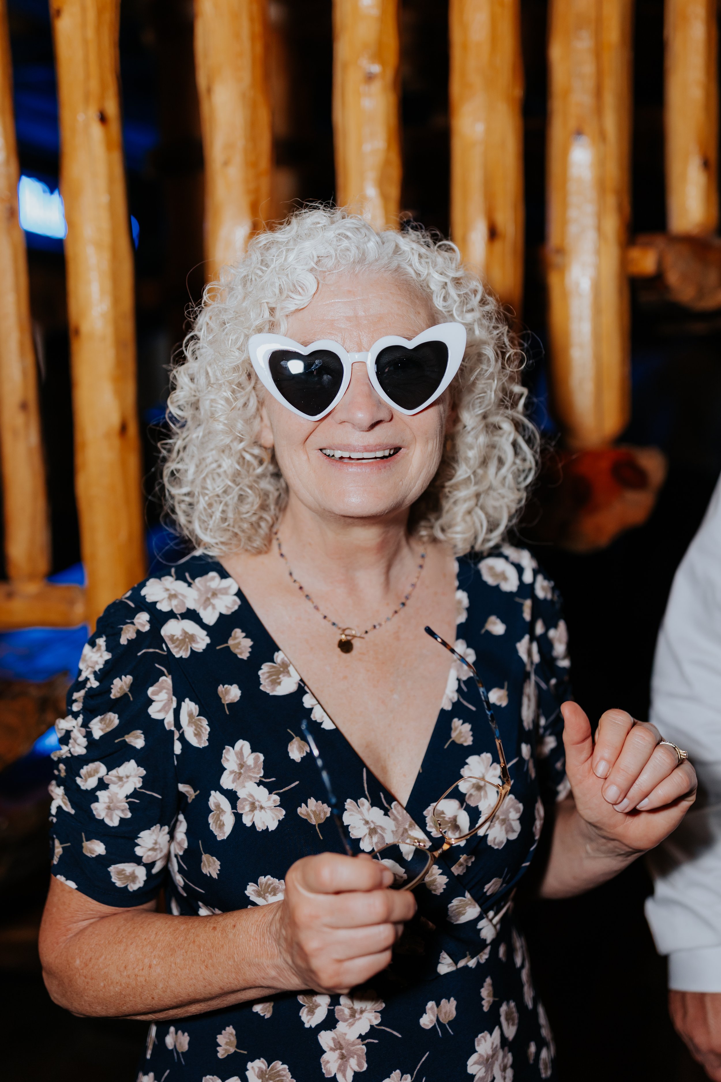 An elderly woman with curly blonde hair wearing oversized heart-shaped sunglasses, a floral dress, and holding glasses, smiling at the camera.