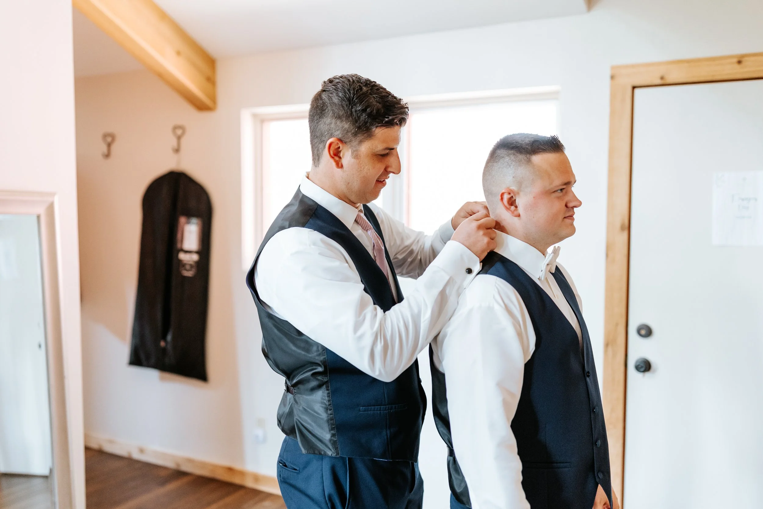 Two men in formal vests and white shirts preparing for a formal event, with one helping the other adjust his collar in a well-lit room.