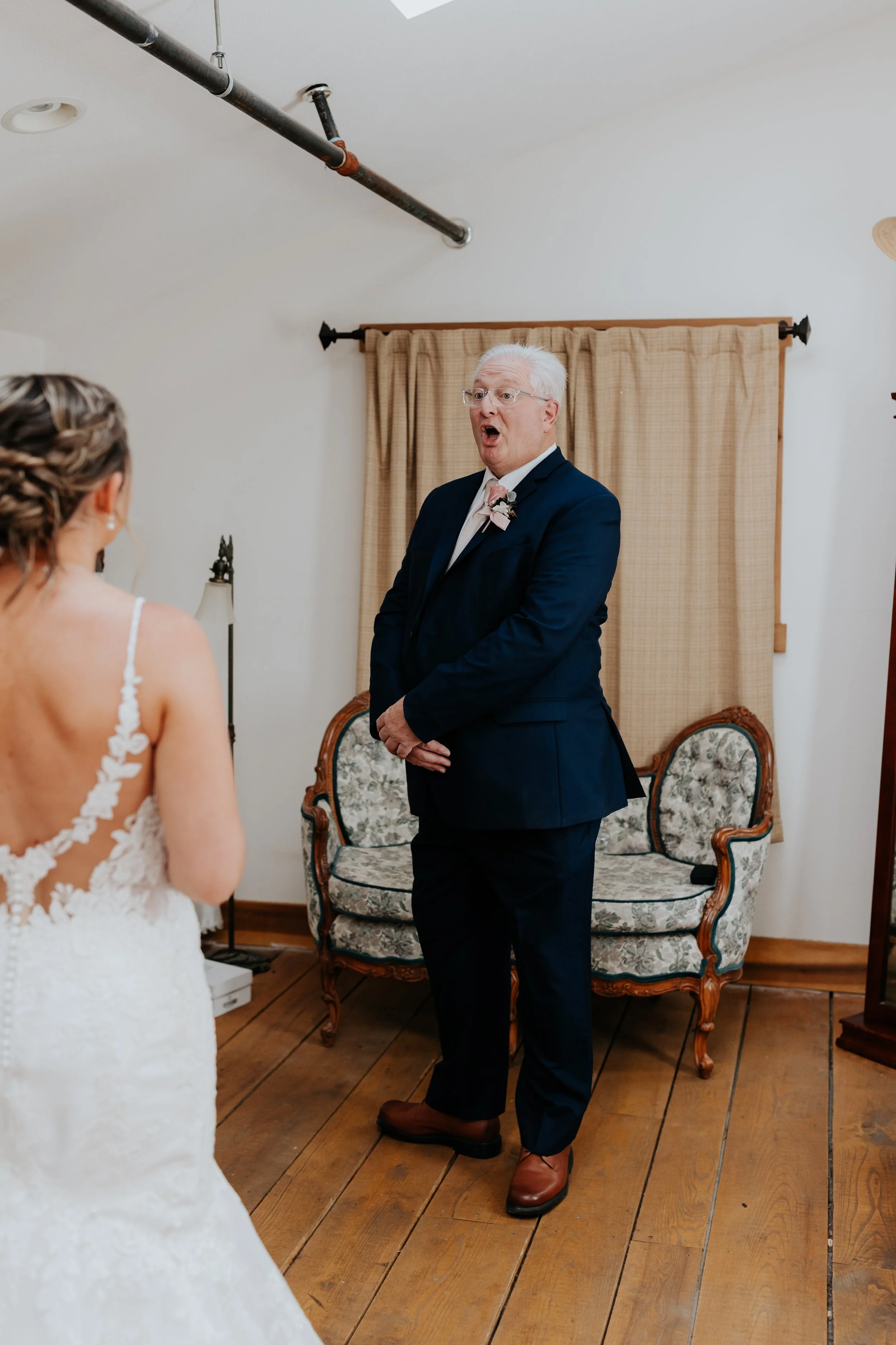An elderly man in a navy blue suit with a white boutonniere is surprise or emotional, standing in front of a bride in a wedding dress, inside a room with wooden floor and beige curtains.