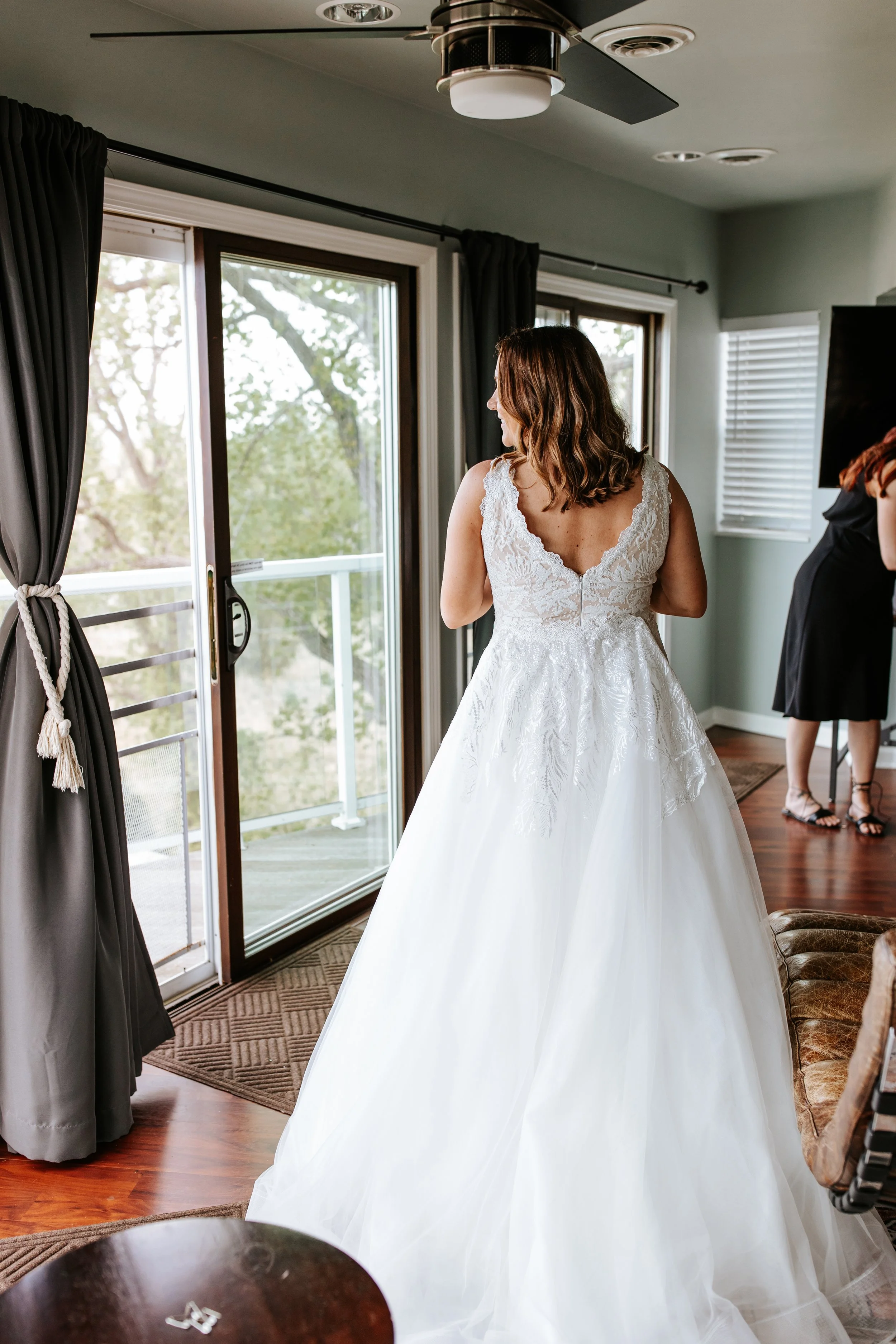 A woman in a white wedding dress standing by an open sliding glass door, looking outside. Another woman in black is in the background near a television.
