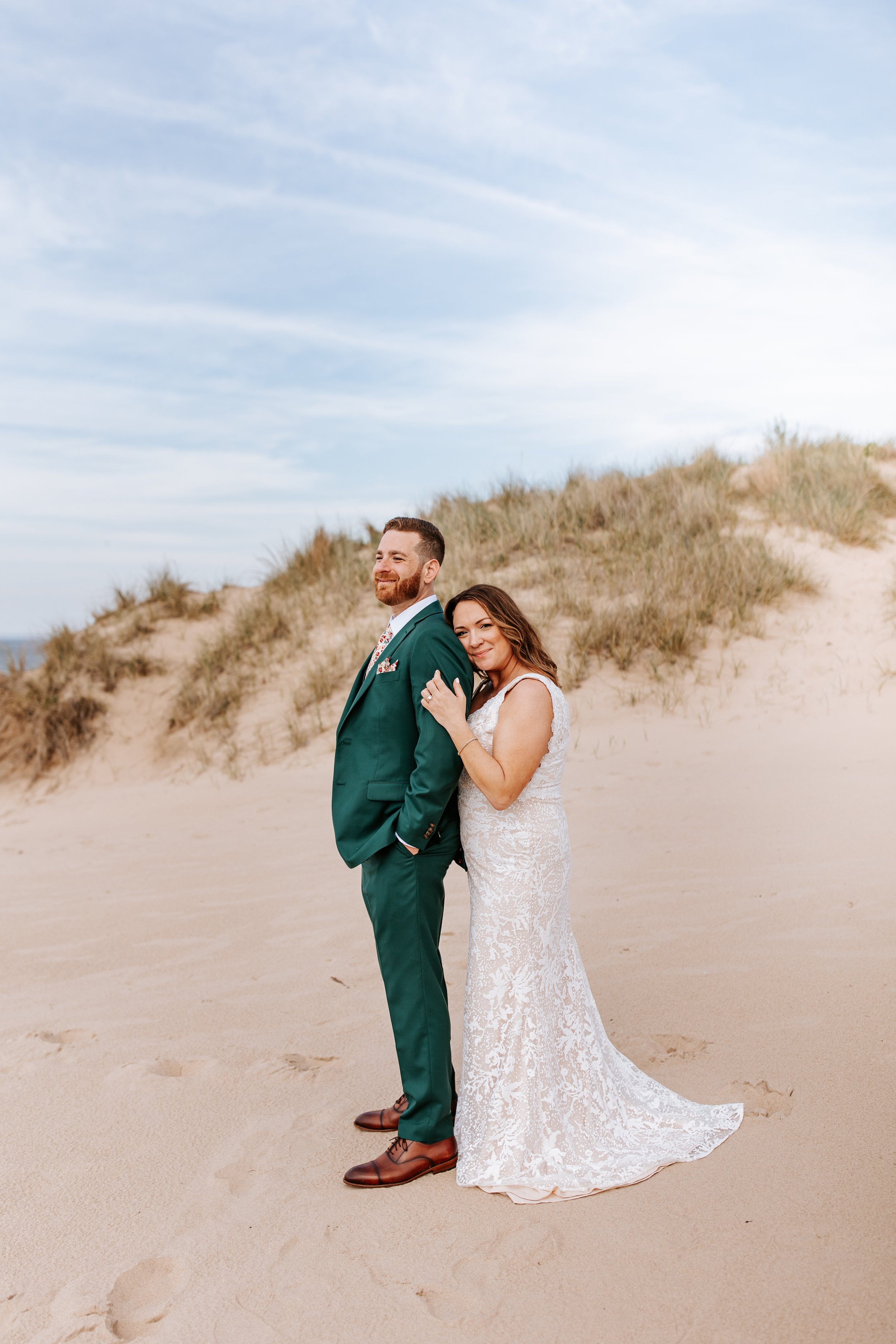 Bride and groom standing on a sandy beach with grassy dunes in the background, smiling and leaning on each other.