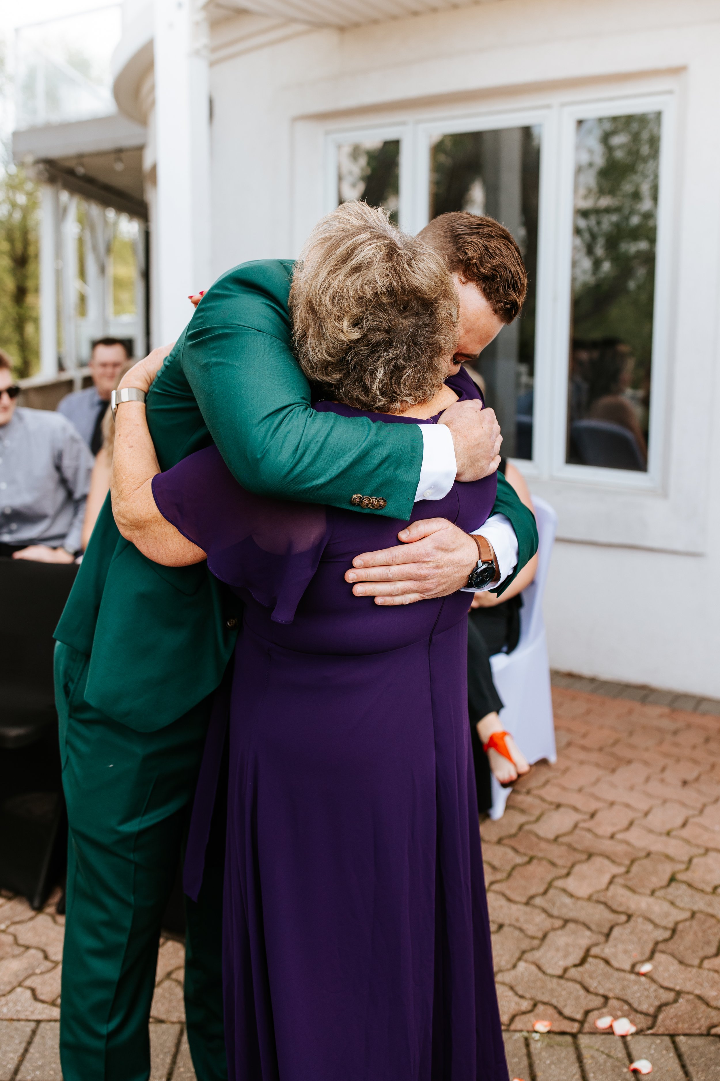 A man and an elderly woman hugging warmly at an outdoor event, with other people sitting and standing in the background.