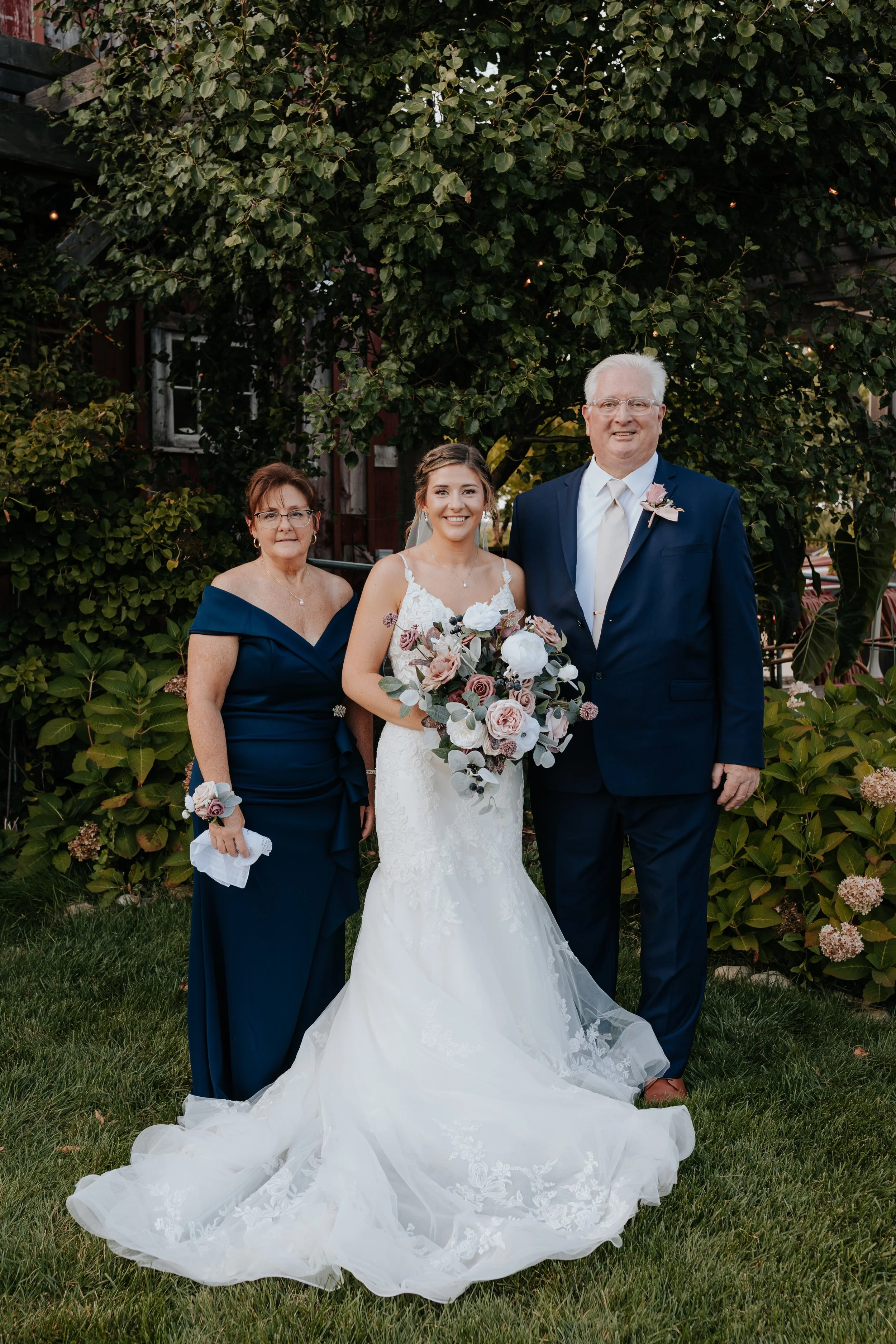 A bride in a white wedding dress holding a bouquet, standing between an older woman in a navy blue dress and an older man in a navy suit, outdoors with greenery and flowering plants.