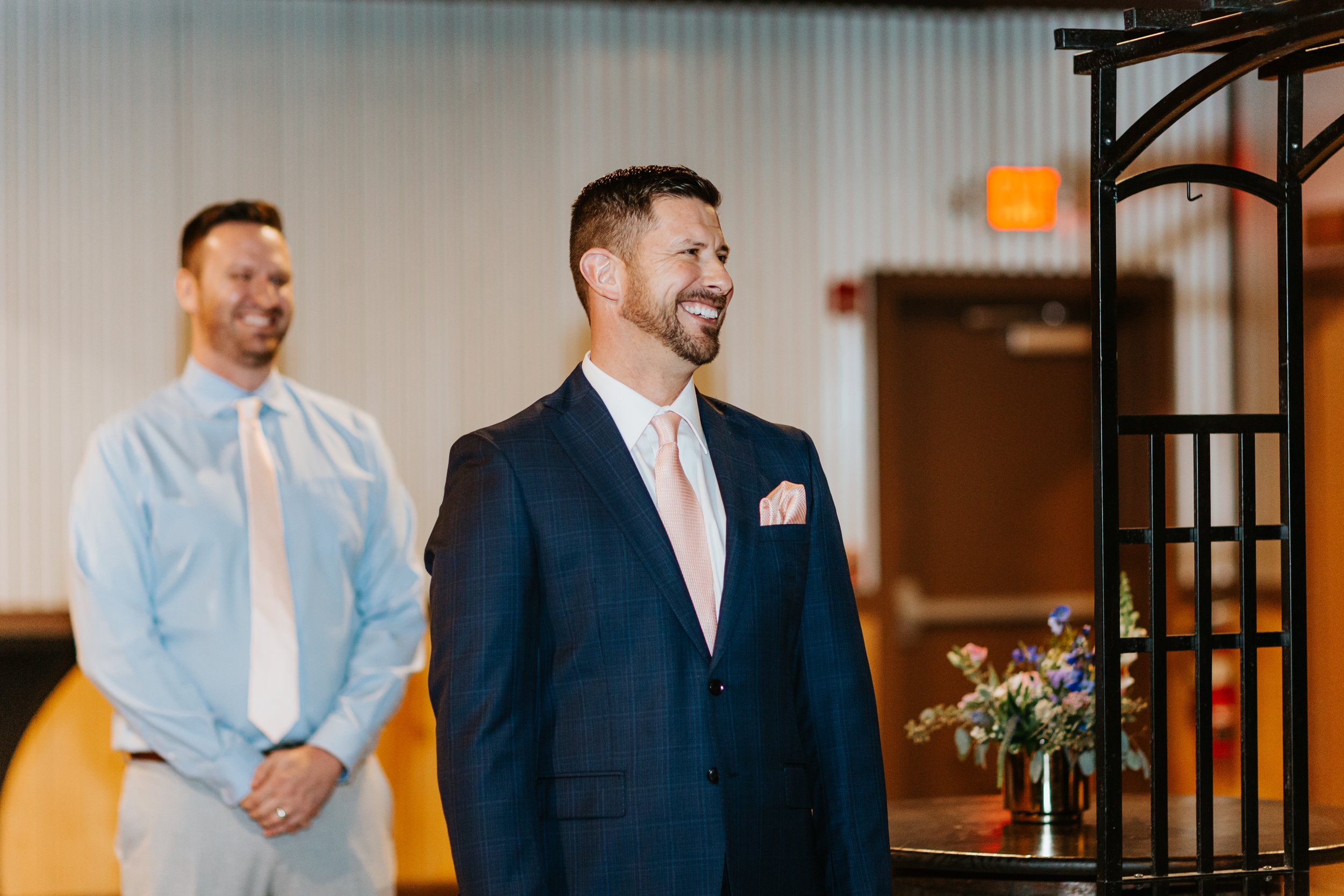 Two men in suits smiling at an indoor event, with a black decorative arch and a flower arrangement nearby.