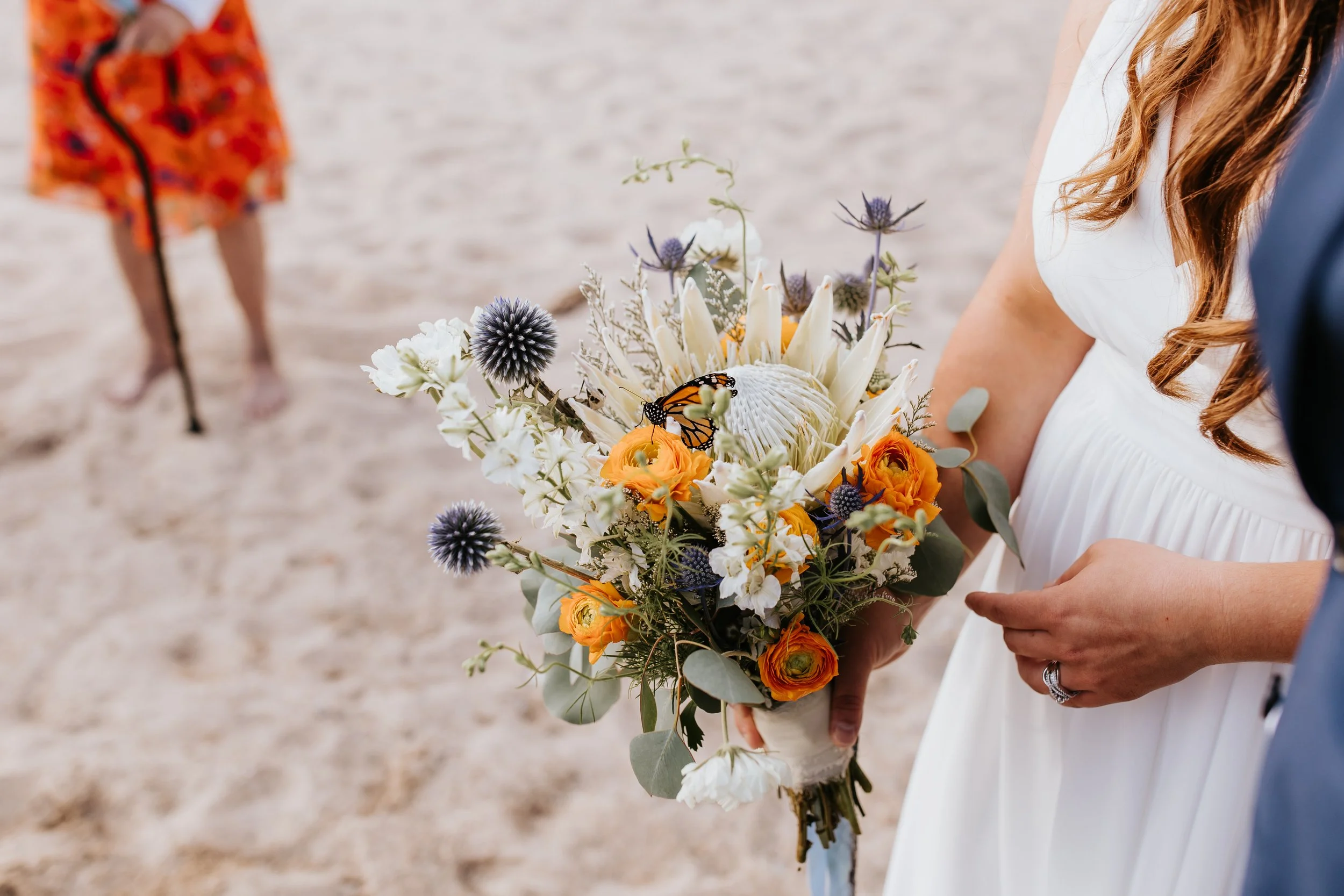 Bride holding a bouquet of orange, white, and purple flowers during a beach wedding.