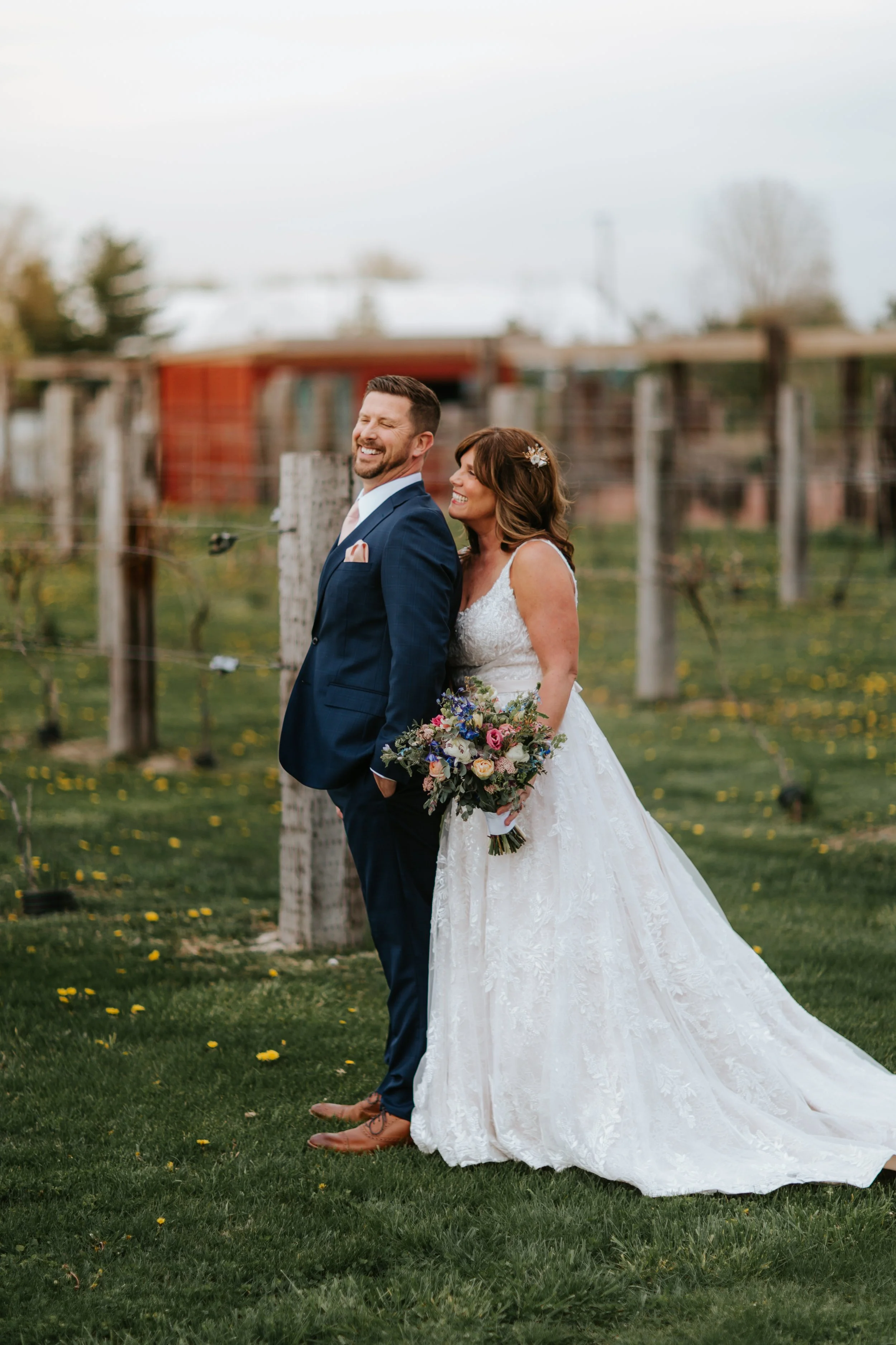 A bride and groom in wedding attire standing outdoors on a grassy field, smiling and laughing. The bride is holding a bouquet of flowers, and the groom has his hands in his pockets. There are barn and tree structures in the background.